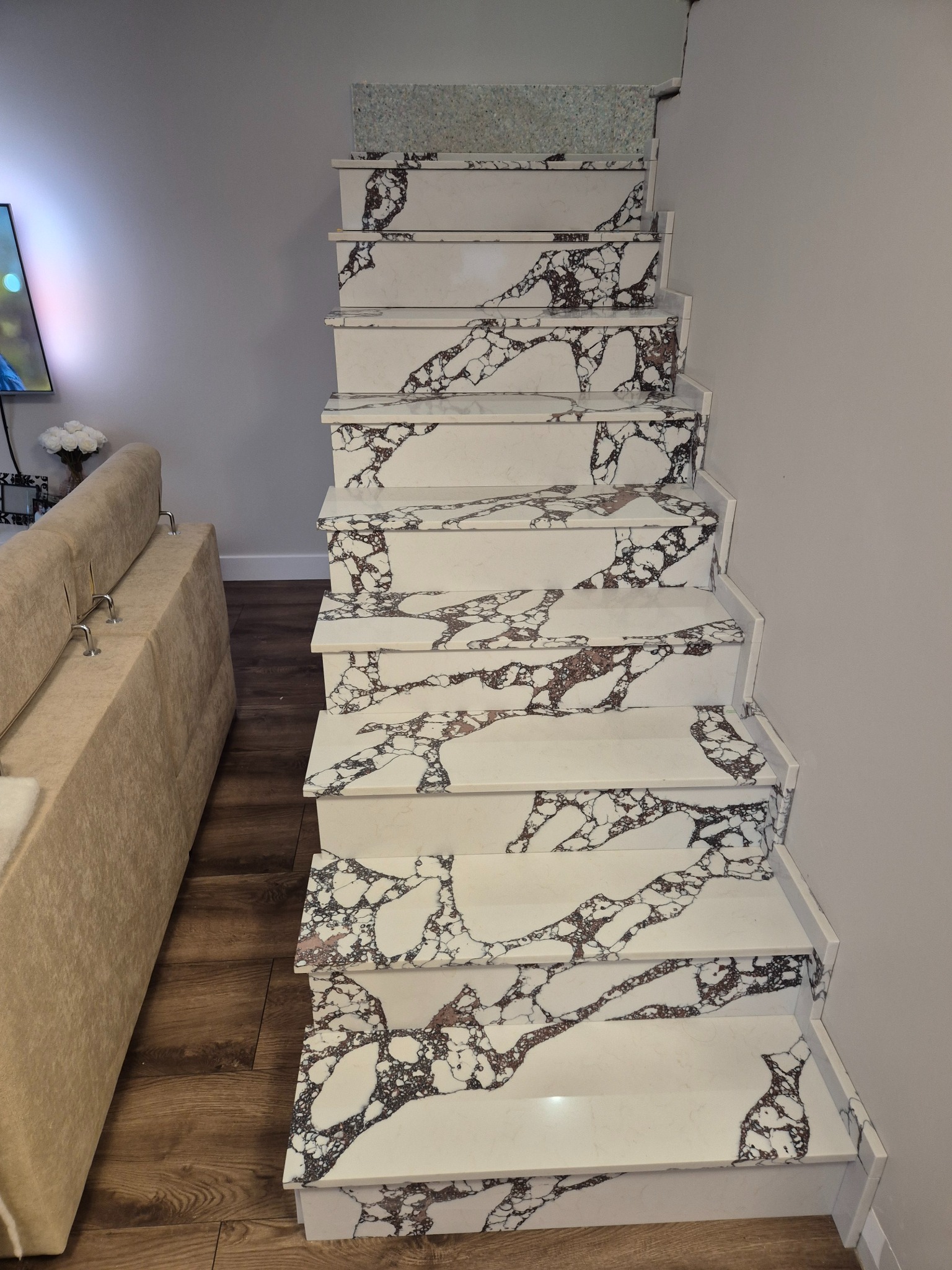 Schody z białego kamienia z unikalnym wzorem żyłkowania w odcieniach brązu i czerni, widok z góry, z fragmentem sofy w tle. Eleganckie wykończenie wnętrza.