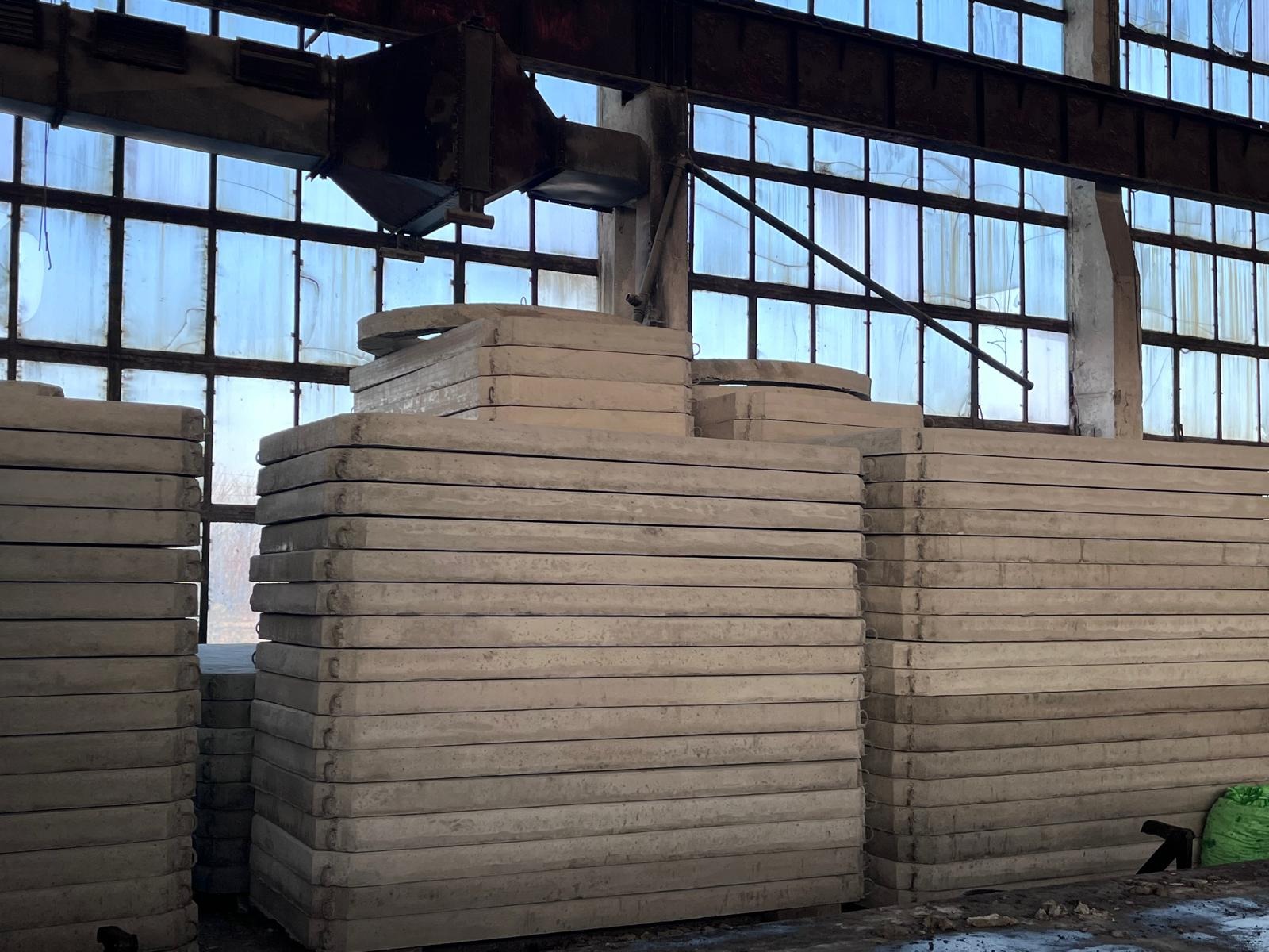 Stosy płyt betonowych z uchwytami, składowane w hali w Radymnie, widoczne okna i konstrukcja stalowa pod sufitem.