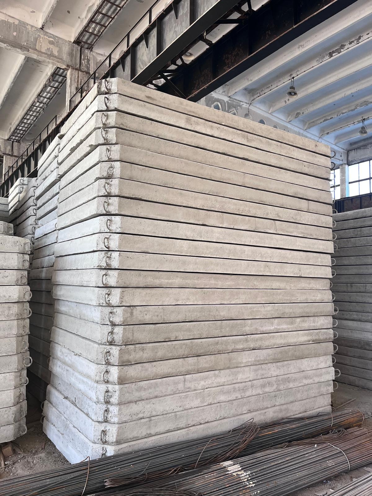 Stos szarych, betonowych płyt z metalowymi uchwytami w magazynie, obok leżą wiązki prętów zbrojeniowych.