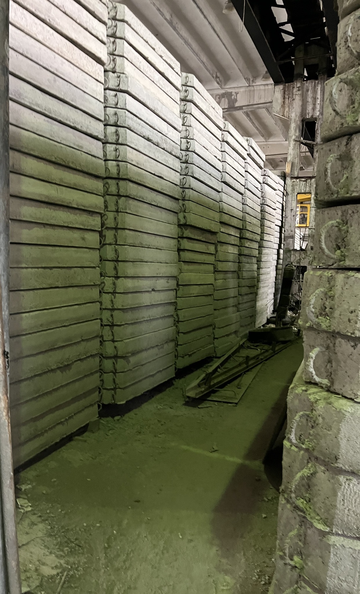 Magazyn prefabrykatów betonowych: stosy szarych płyt z metalowymi uchwytami, oświetlenie przemysłowe, surowy betonowy podkład.