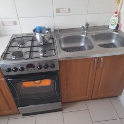 Darek'S NEXUS Dariusz Szmalc - Kuchenny zlew dwukomorowy z drewnianymi szafkami poniżej, obok kuchenka gazowa z garnkiem i piekarnikiem w trakcie pieczenia, białe kafelki na ścianie i podłodze.