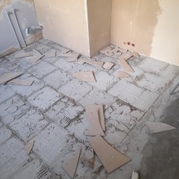 Darek'S NEXUS Dariusz Szmalc - Demontaż starej podłogi z płytek w rogu pomieszczenia, widoczne fragmenty płytek i resztki kleju, ściany w kolorze beżowym i zielonym, instalacja wodna.