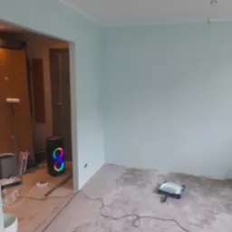 Świeżo pomalowane na jasnozielono ściany pokoju, widoczny wałek i kuweta malarska na podłodze oraz głośnik z kolorowym podświetleniem w przejściu do drugiego pomieszczenia.