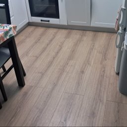 Darek'S NEXUS Dariusz Szmalc - Jasne panele podłogowe w kuchni z widocznym fragmentem stołu z kolorowym obrusem, białymi szafkami i lodówką.