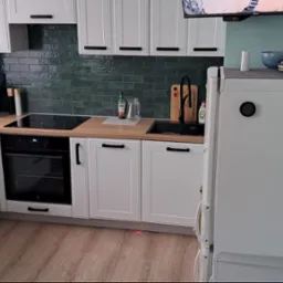 Nowoczesna kuchnia z białymi szafkami, drewnianym blatem, ciemnozielonymi płytkami na ścianie, czarną armaturą i sprzętami AGD, w tym piekarnikiem Electrolux i lod&oacute;wką z magnesami.