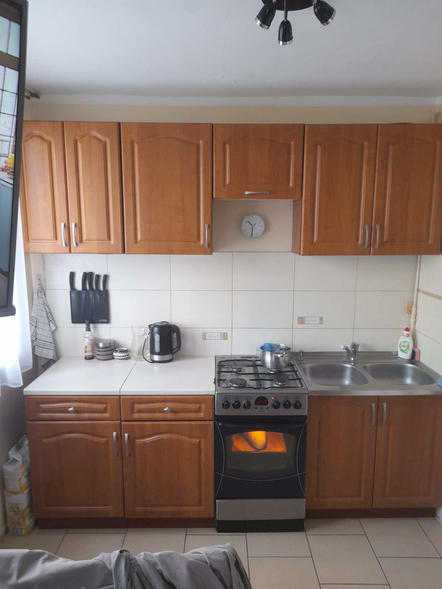 Kuchnia z drewnianymi szafkami, białą glazurą, kuchenką gazową z piekarnikiem i dwukomorowym zlewem, widok na ścianę z wyposażeniem.