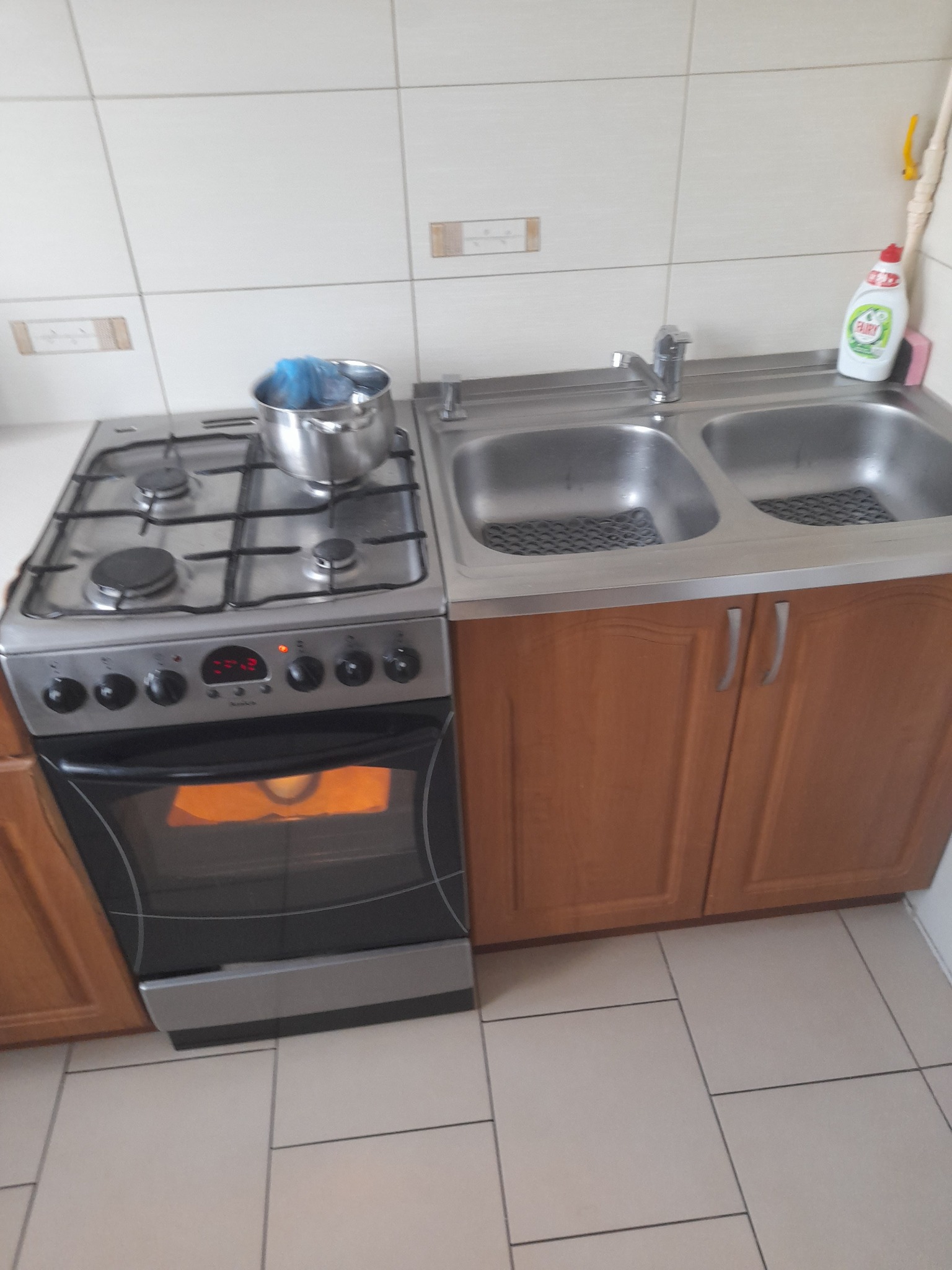 Kuchenny zlew dwukomorowy z drewnianymi szafkami poniżej, obok kuchenka gazowa z garnkiem i piekarnikiem w trakcie pieczenia, białe kafelki na ścianie i podłodze.