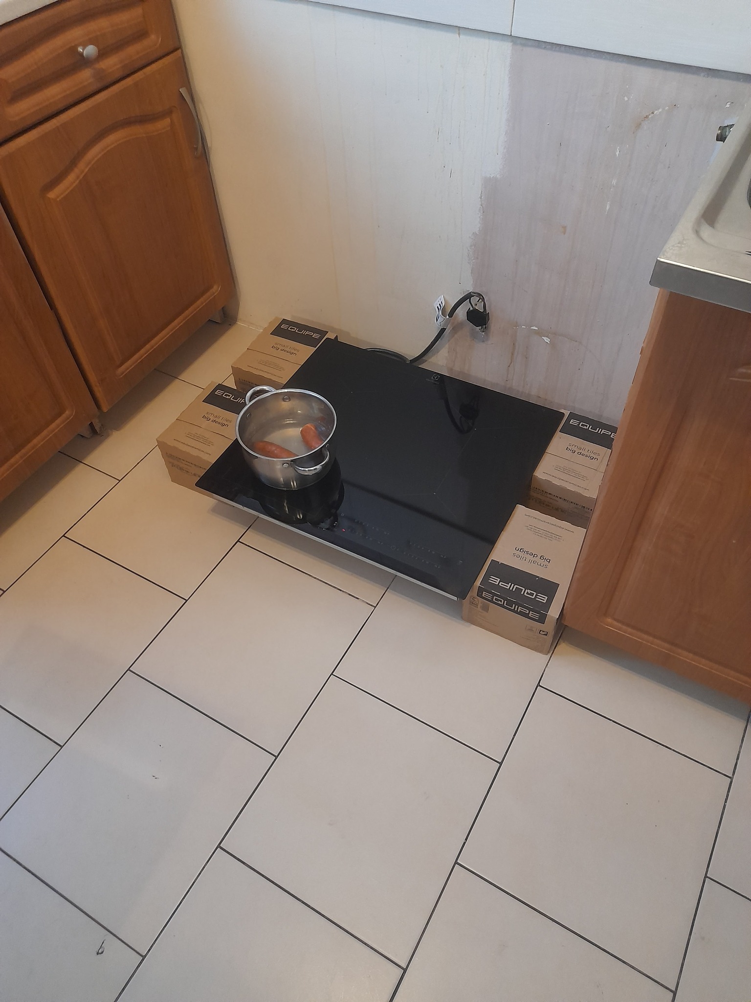 Nowa, czarna płyta indukcyjna, częściowo podparta kartonami producenta płytek podłogowych, z garnkiem i gotującymi się parówkami, ustawiona na podłodze z białych płytek, obok niedokończonej ściany...