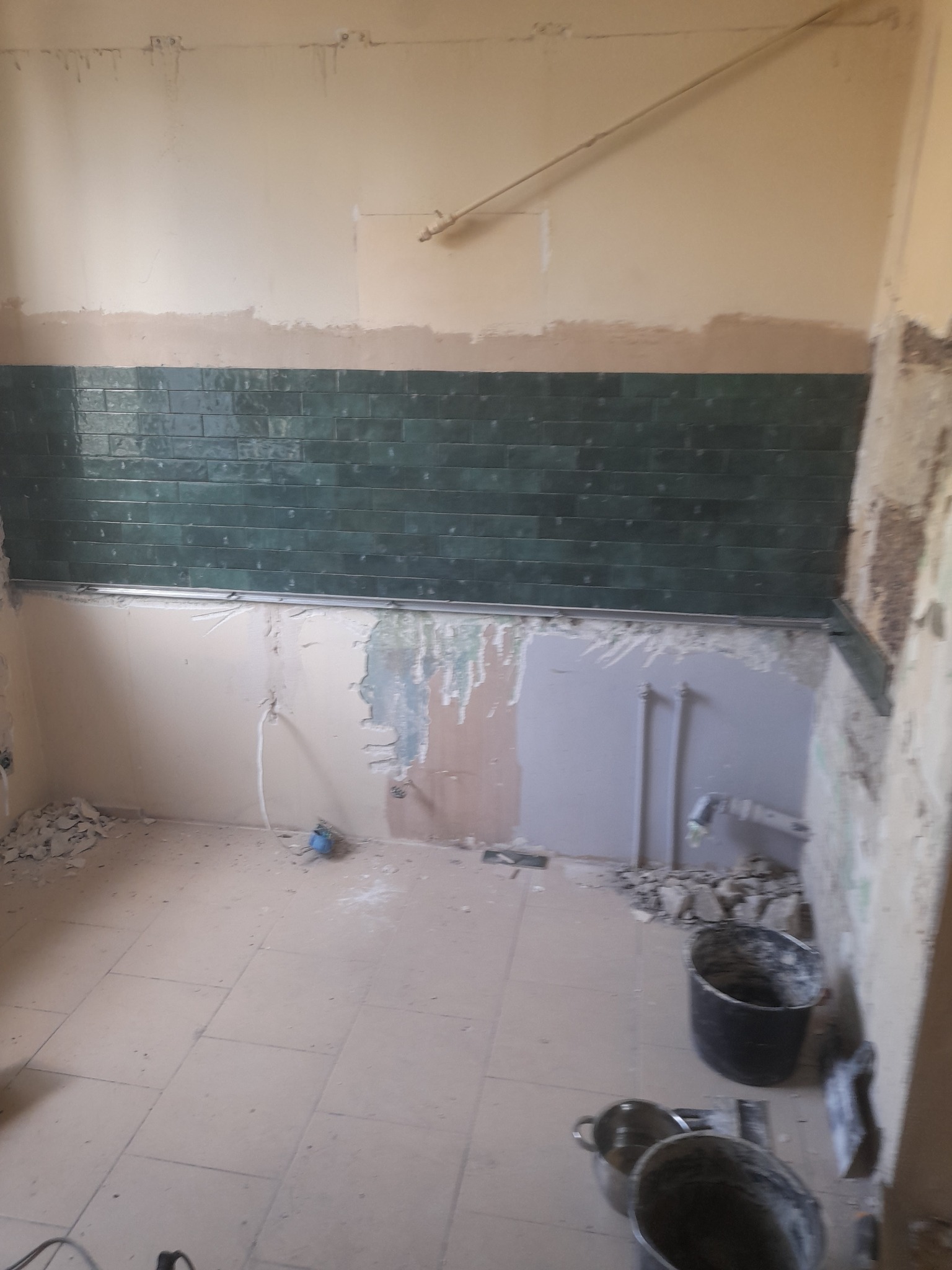 Fragment ściany z częściowo położonymi ciemnozielonymi płytkami, widoczne ślady po skuciu starych płytek, instalacja elektryczna i hydrauliczna, dwa wiadra i szpachelka na podłodze z płytek