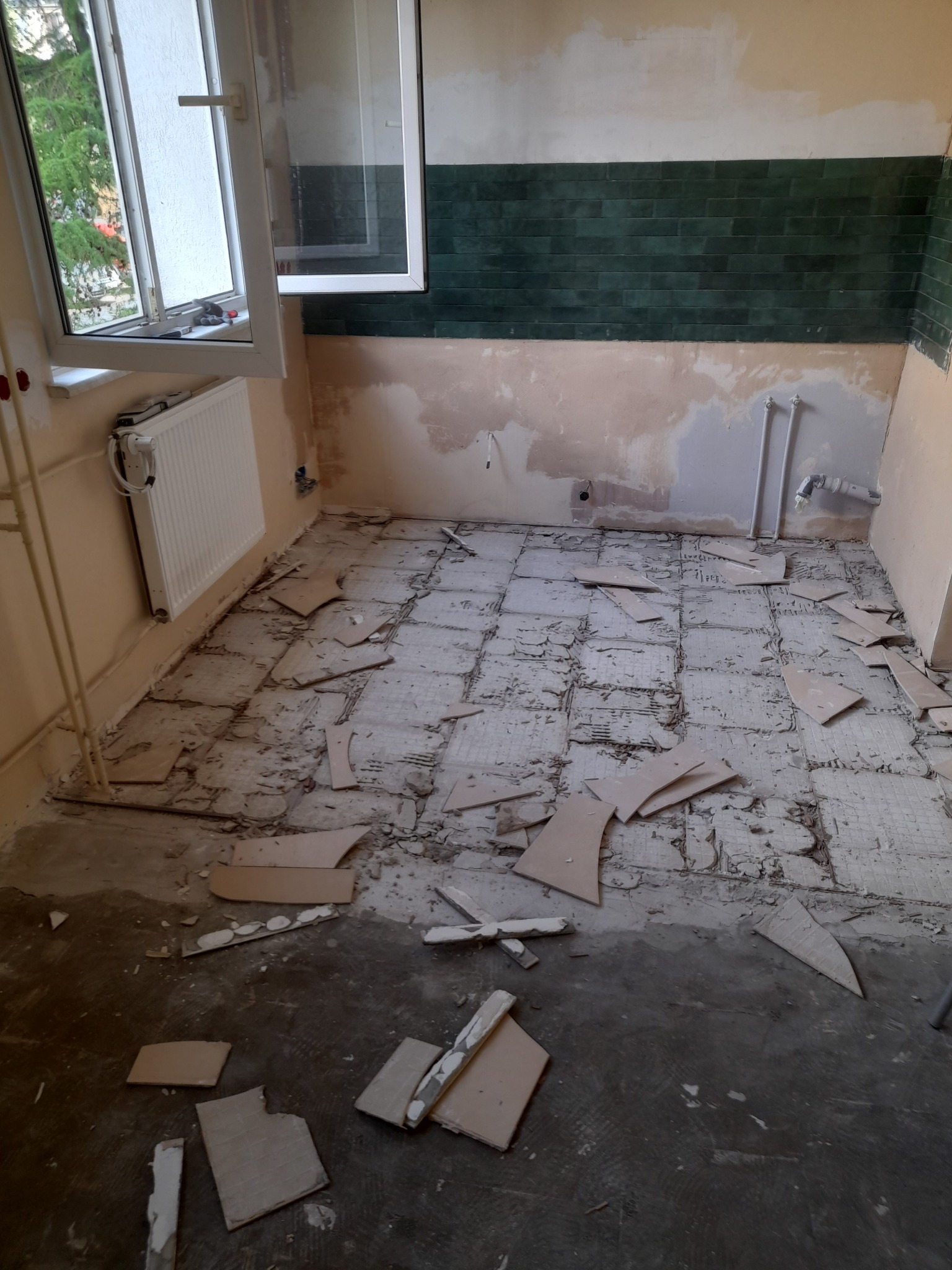 Demontaż starej podłogi w trakcie remontu kuchni, widoczne fragmenty płytek i resztki kleju na posadzce, ściany częściowo przygotowane pod nowe wykończenie, okno otwarte.