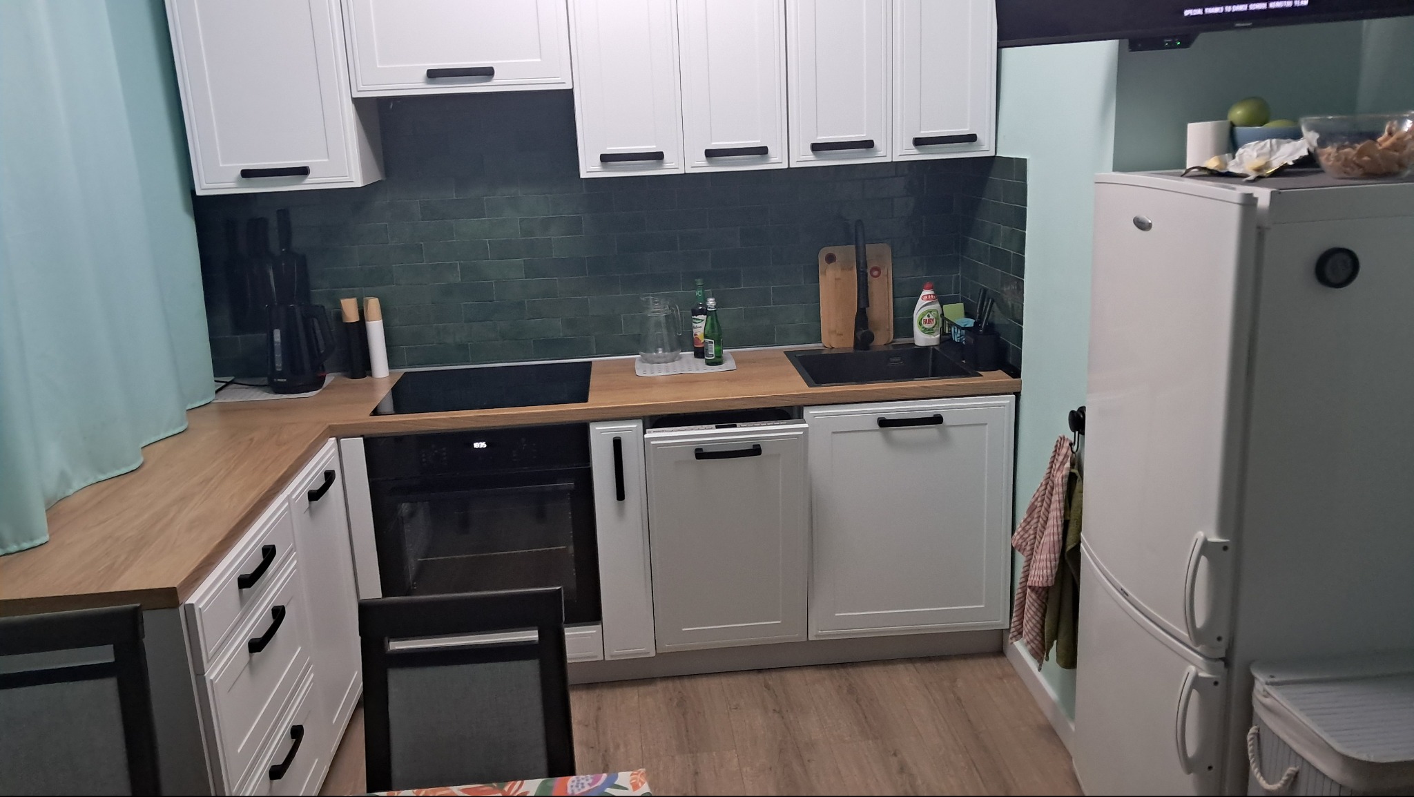 Nowoczesna kuchnia z białymi szafkami z czarnymi uchwytami, drewnianym blatem, zielonymi płytkami na ścianie i czarnym zlewem.