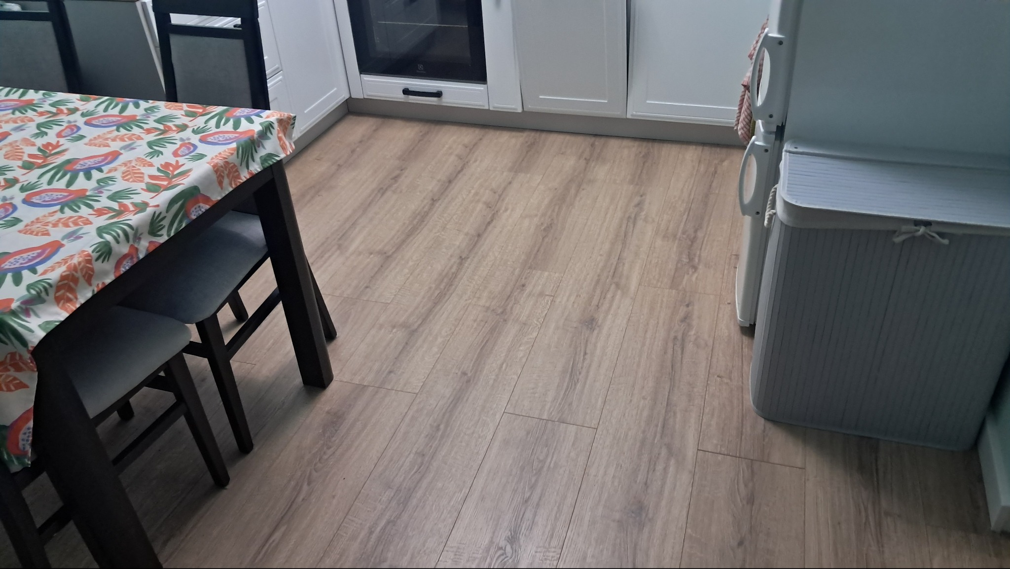 Jasne panele podłogowe w kuchni z widocznym fragmentem stołu z kolorowym obrusem, białymi szafkami i lodówką.