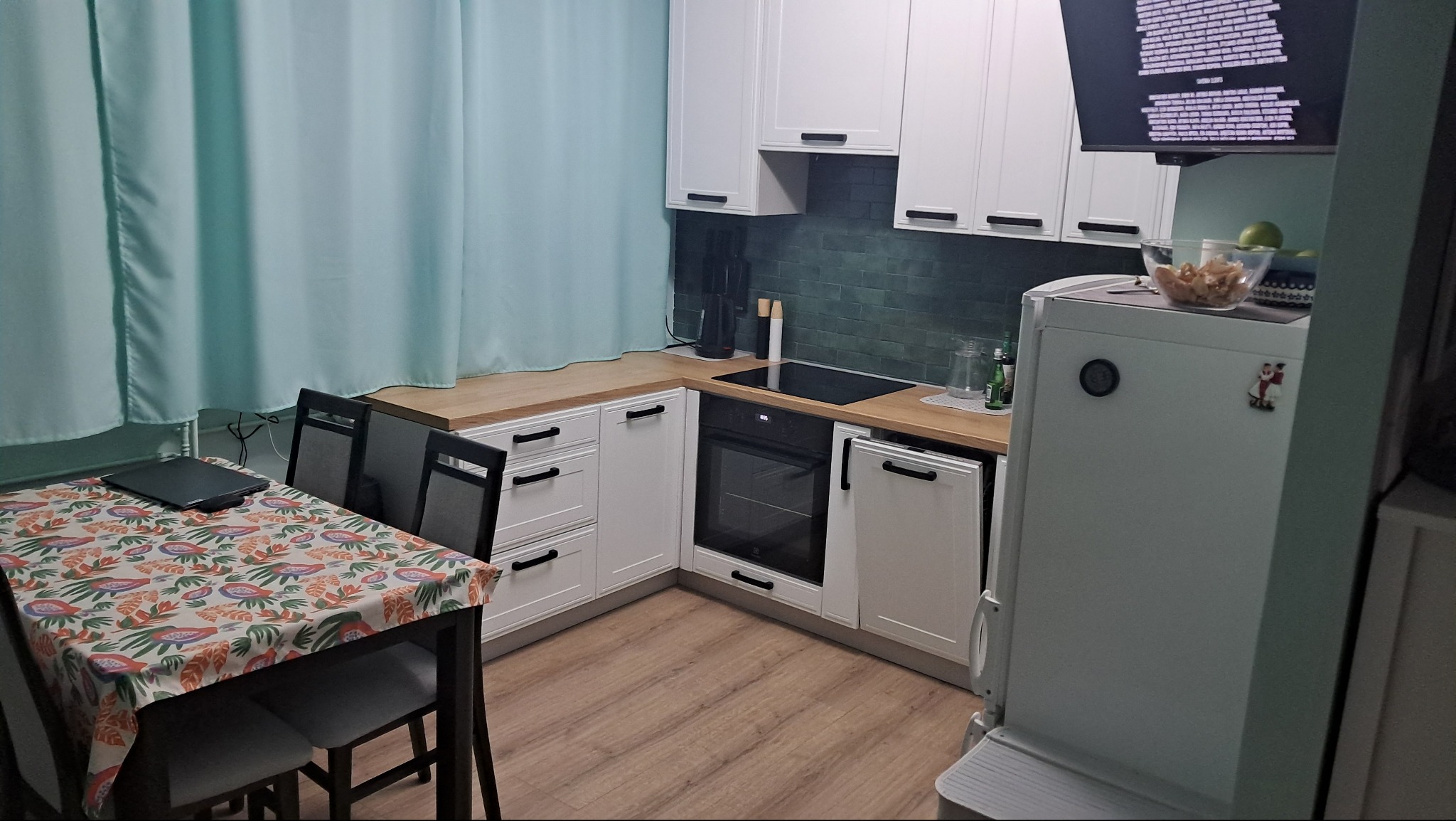 Nowoczesna kuchnia z białymi szafkami, drewnianym blatem, zielonymi płytkami na ścianie i stołem z kolorowym obrusem, oświetlona naturalnym światłem