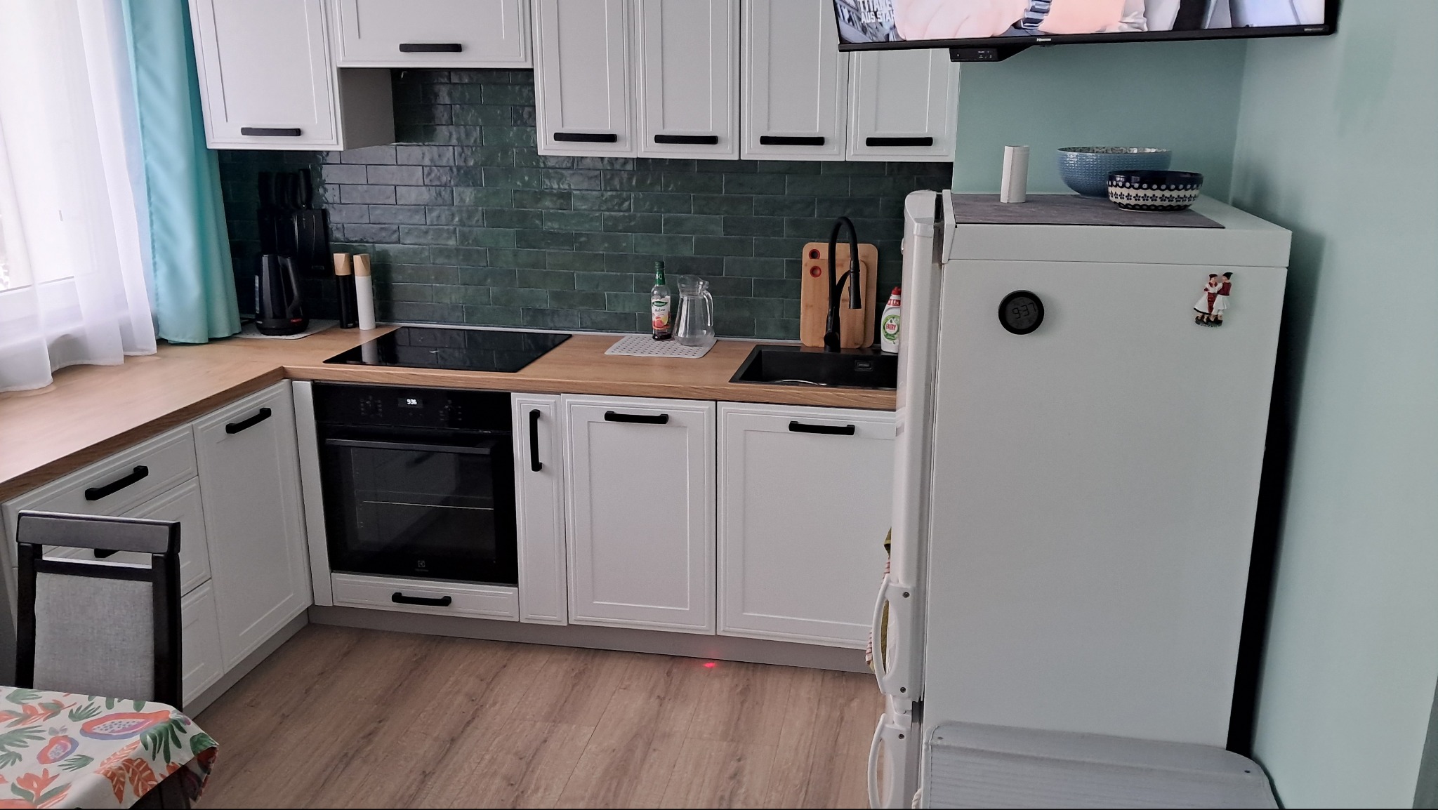 Nowoczesna kuchnia z białymi szafkami, drewnianym blatem, ciemnozielonymi płytkami na ścianie, czarną armaturą i sprzętami AGD, w tym piekarnikiem Electrolux i lodówką z magnesami.