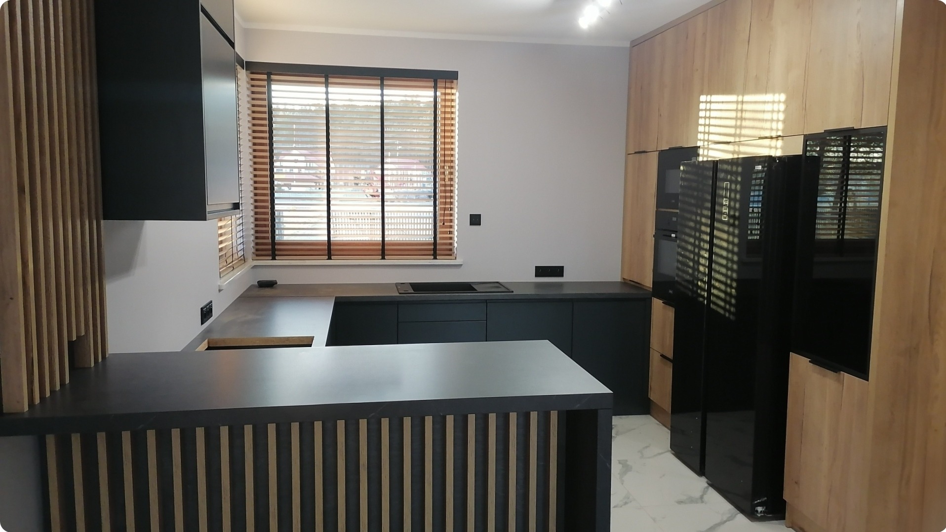 Nowoczesna kuchnia z czarnymi szafkami i blatem, drewnianymi lamelami na wyspie oraz drewnianymi żaluzjami w oknie.