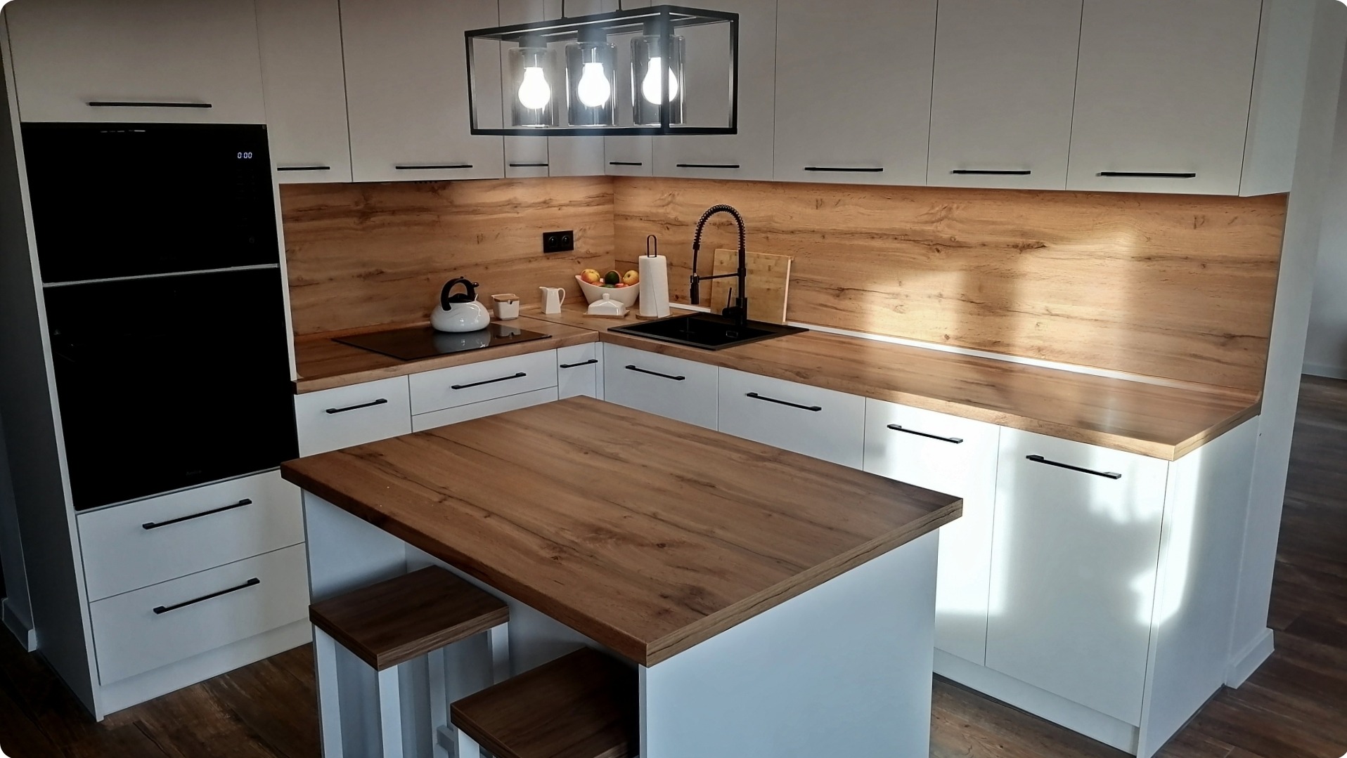Nowoczesna kuchnia z białymi szafkami, drewnianym blatem i wyspą z dwoma stołkami, oświetlona lampą z trzema żarówkami w czarnej obudowie.