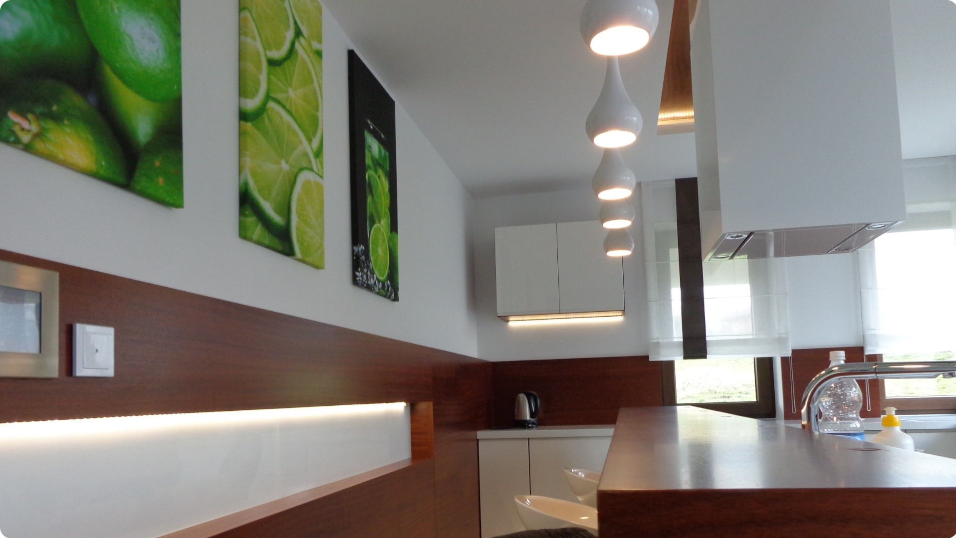 Nowoczesna kuchnia z drewnianym blatem, białymi szafkami i wiszącymi lampami. Na ścianie grafiki z motywem limonki.