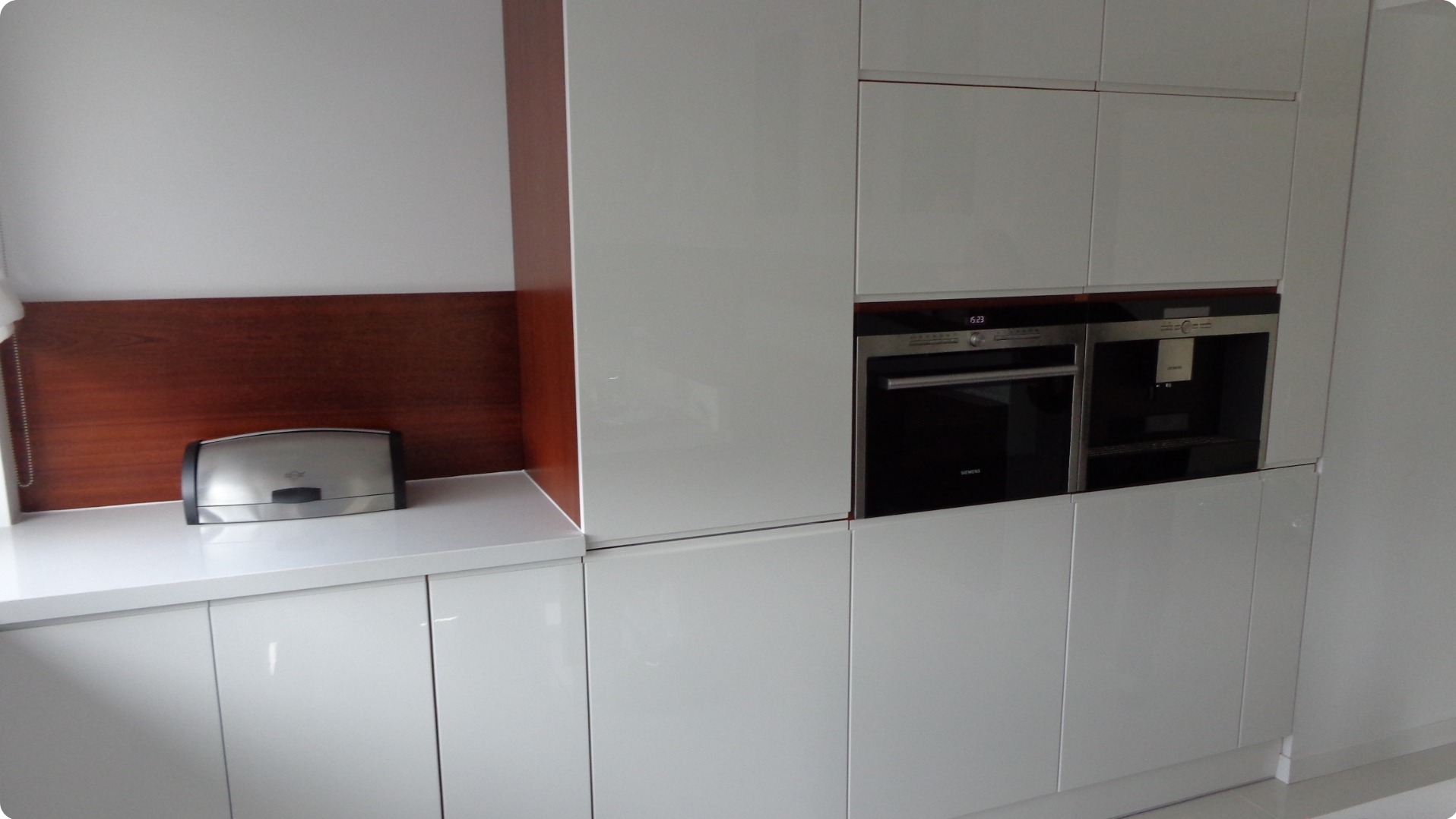 Biała, nowoczesna zabudowa kuchenna z piekarnikiem i ekspresem do kawy w zabudowie, uzupełniona o drewniane wykończenie i metalowy chlebak.