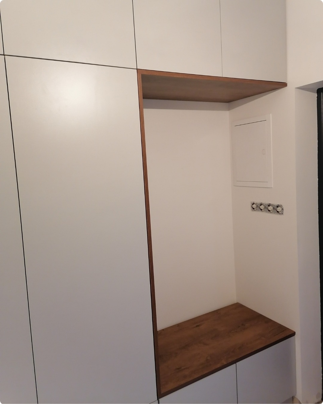 Wnęka z drewnianą półką i siedziskiem w zabudowie szafy z białymi frontami, widoczne gniazdka elektryczne na ścianie.