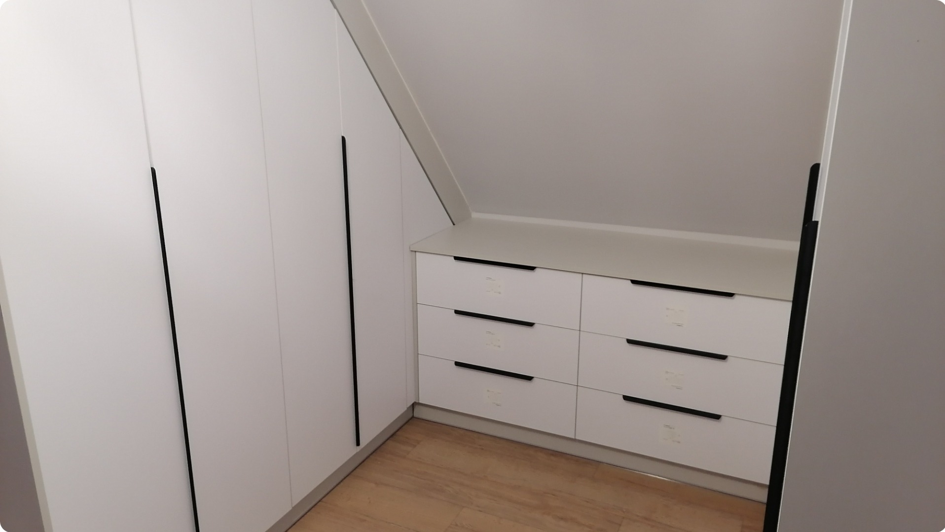 Biała szafa wnękowa z sześcioma szufladami pod skosem dachu, z minimalistycznymi czarnymi uchwytami, na podłodze panele imitujące drewno.