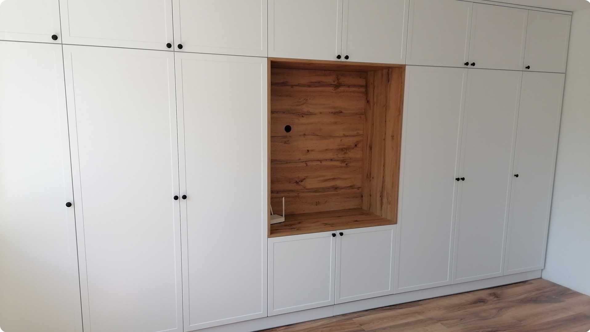 Biała zabudowa meblowa z wnęką wykończoną drewnopodobnym dekorem, widoczny router i otwór na kable, podłoga drewniana.
