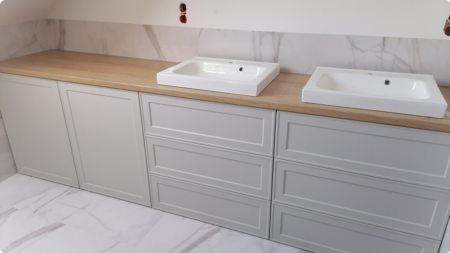Jasnoszare szafki łazienkowe z drewnianym blatem i dwiema białymi umywalkami nablatowymi na tle marmurowych płytek ściennych i podłogowych.