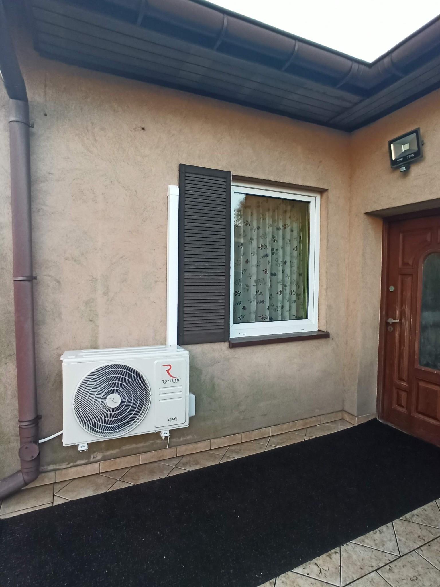 Zewnętrzna jednostka klimatyzacji Rotenso Mirai zamontowana na elewacji domu obok okna z brązową okiennicą.