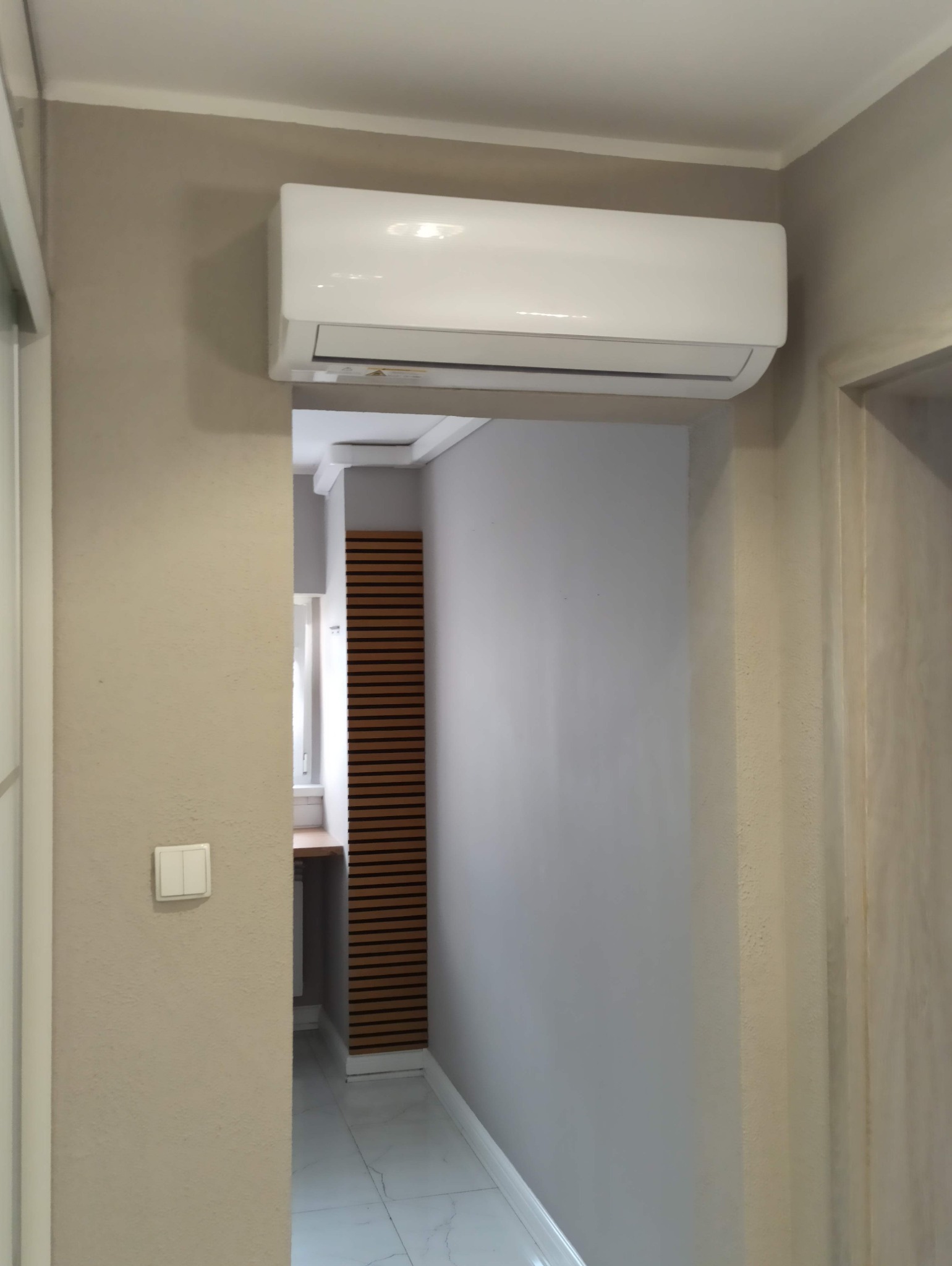 Biały klimatyzator ścienny zamontowany nad wejściem do pomieszczenia z drewnianą dekoracją ścienną z poziomych listew. Widoczne białe ściany i marmurowa podłoga.