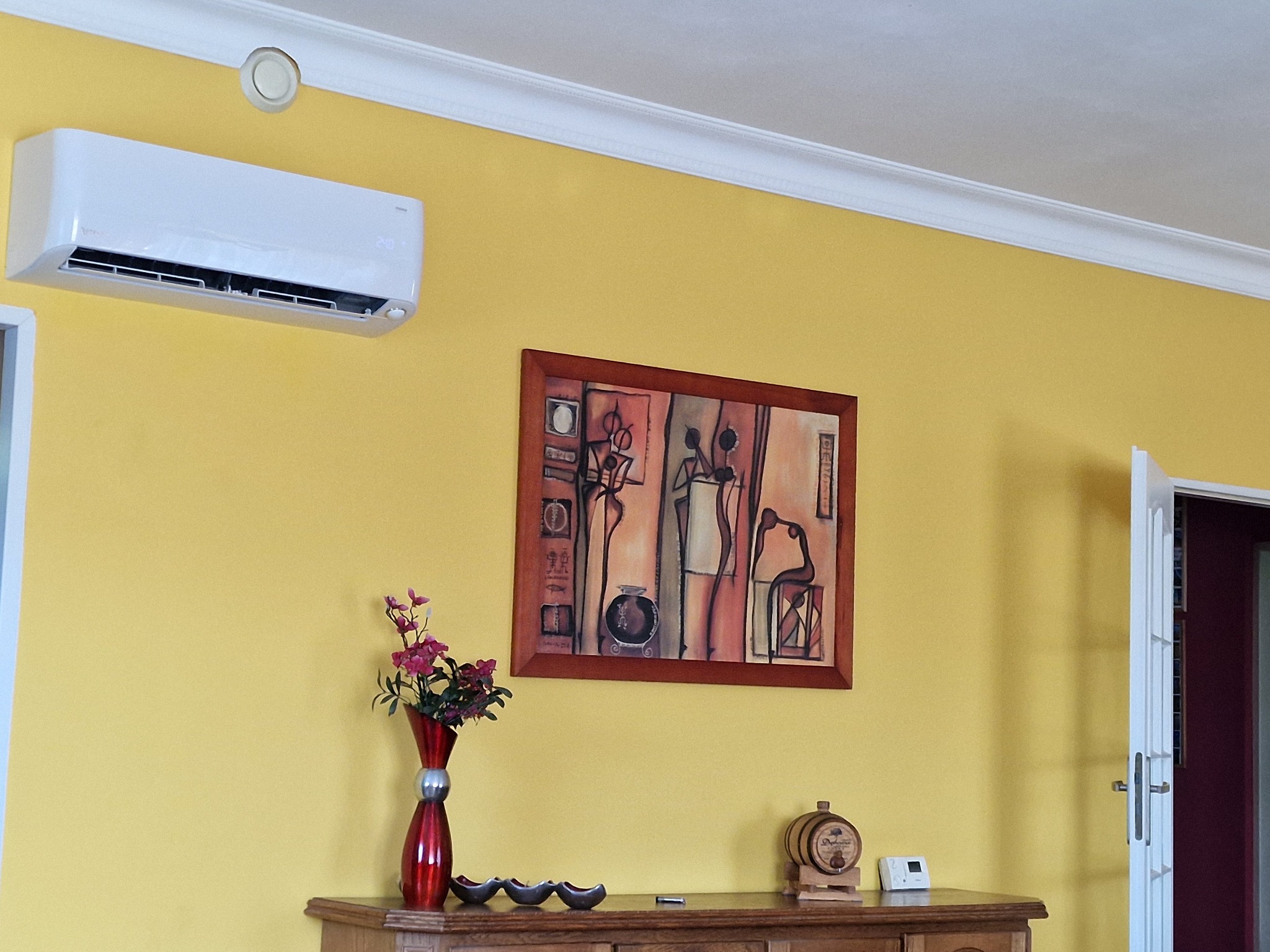 Biały klimatyzator Rotenso Mirai zamontowany na żółtej ścianie w salonie, pod sufitem z dekoracyjnym gzymsem, obok obrazu w brązowej ramie i komody z wazonem kwiatów.