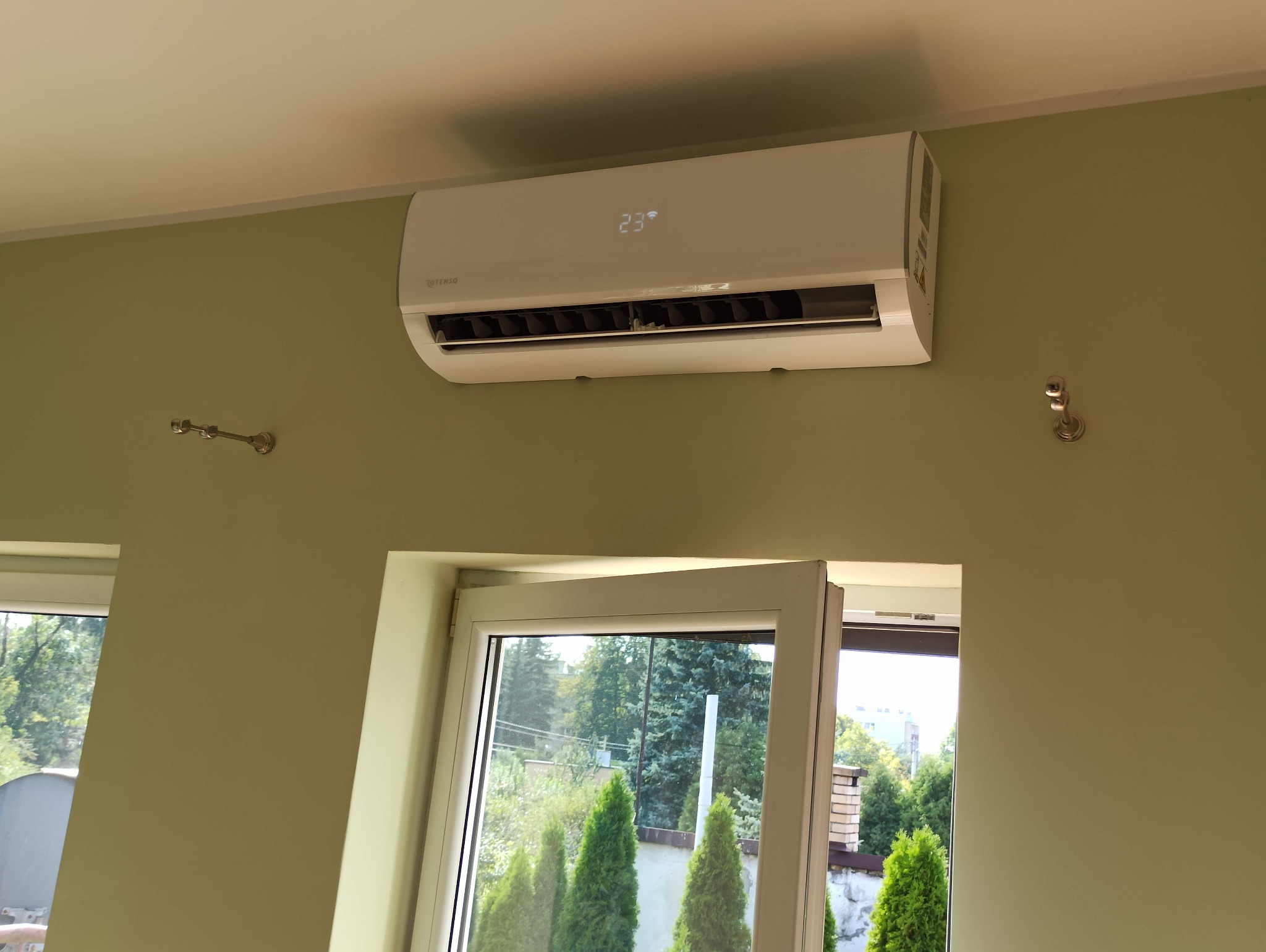 Biały klimatyzator Rotenso Ukura zamontowany na ścianie w pomieszczeniu z oknem, wyświetlający temperaturę 23 stopnie Celsjusza.