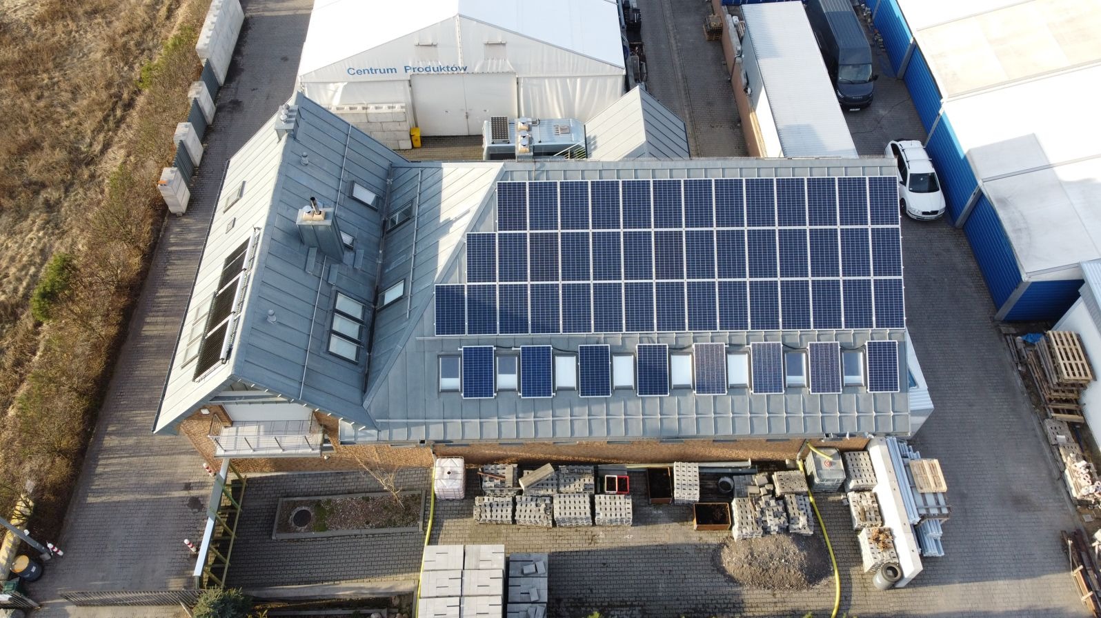 Widok z góry na budynek z szarym dachem pokrytym panelami słonecznymi, obok porozrzucane materiały budowlane, widoczne również sąsiednie budynki i pojazdy.