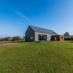 Nowoczesny dom jednorodzinny z elewacją kamienną i dużymi przeszkleniami, otoczony rozległym trawnikiem i naturalnym krajobrazem.