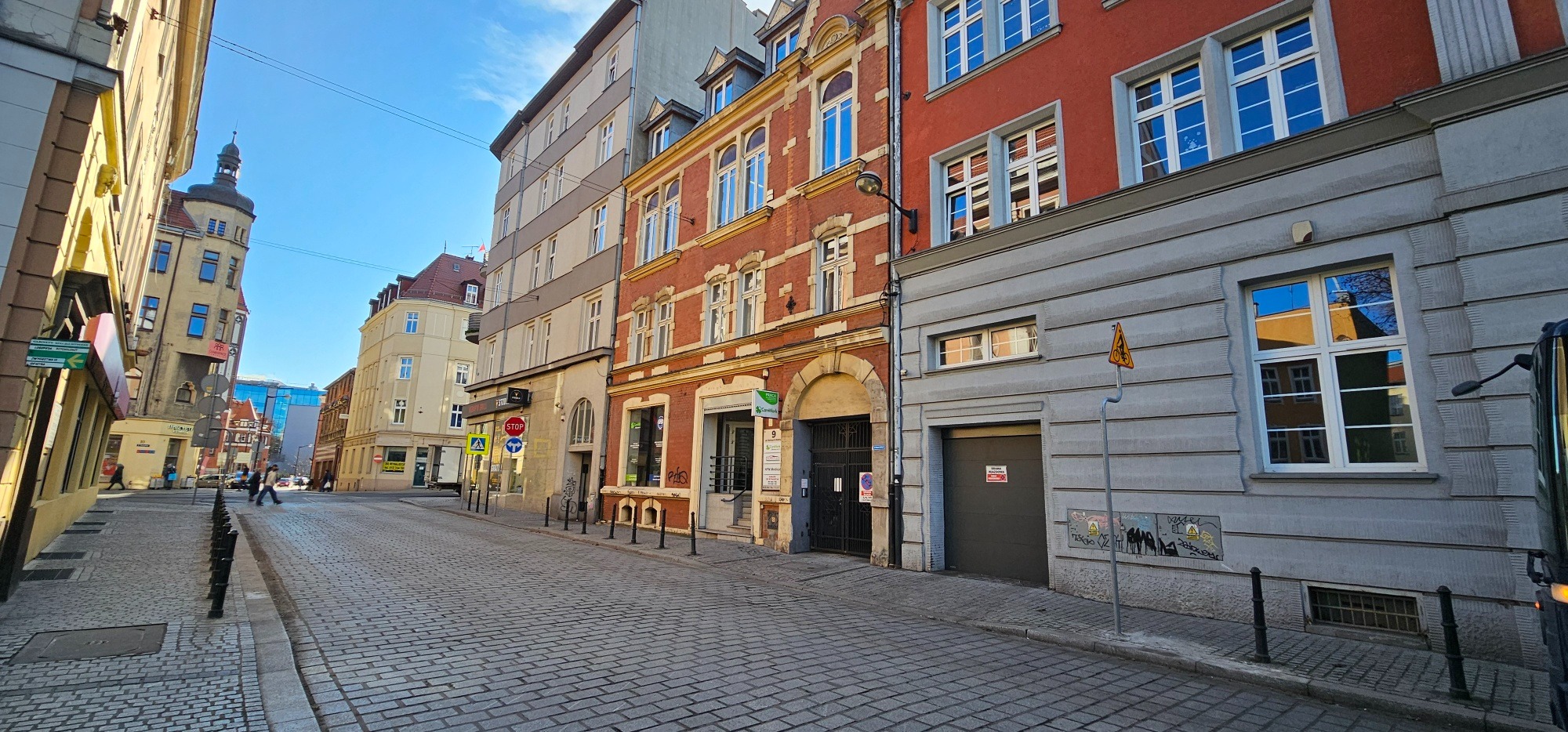 Ulica wybrukowana kostką z budynkami o zróżnicowanej architekturze w słoneczny dzień, widoczne znaki drogowe i elementy małej architektury.