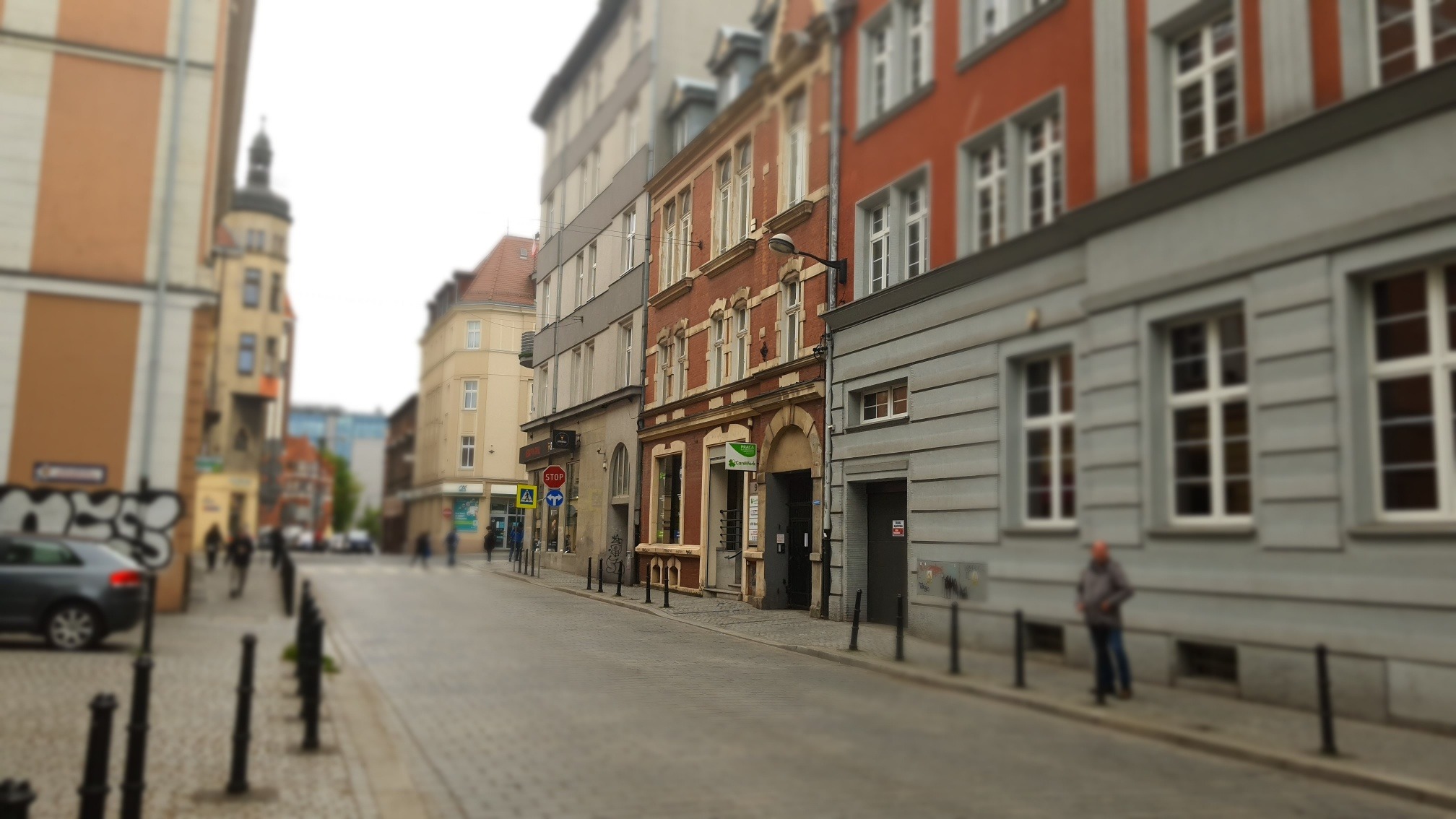 Ulica miejska z budynkami o różnej architekturze, chodnikiem z czarnymi słupkami i przechodniami w oddali, z efektem tilt-shift.