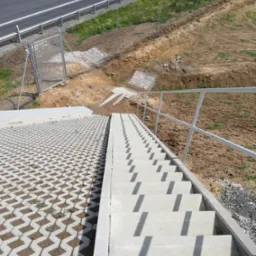 Schody betonowe prowadzące w dół, obok ażurowa nawierzchnia z kostki betonowej, widok z góry na nową drogę i ogrodzenie.