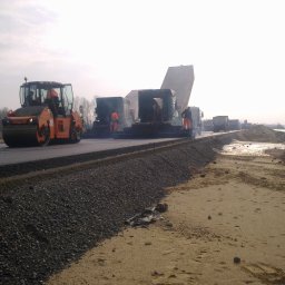 KONSTRUKCJE SILESIA SPÓŁKA Z OGRANICZONĄ ODPOWIEDZIALNOŚCIĄ - Walec drogowy ugniata świeżo położony asfalt na budowie drogi, w tle ekipa robotników obsługuje maszynę rozkładającą nawierzchnię, a za nią ustawia się kolejka ciężarówek z materiałem.