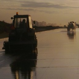 KONSTRUKCJE SILESIA SPÓŁKA Z OGRANICZONĄ ODPOWIEDZIALNOŚCIĄ - Dwa traktory z przyczepami pracują na mokrej, nowo wylanej drodze, odbijając zachodzące słońce.