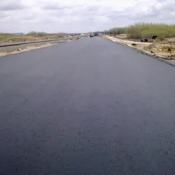 Nowo położona, gładka nawierzchnia asfaltowa drogi w trakcie budowy, z widocznymi maszynami budowlanymi w oddali i elementami przygotowania terenu po bokach.