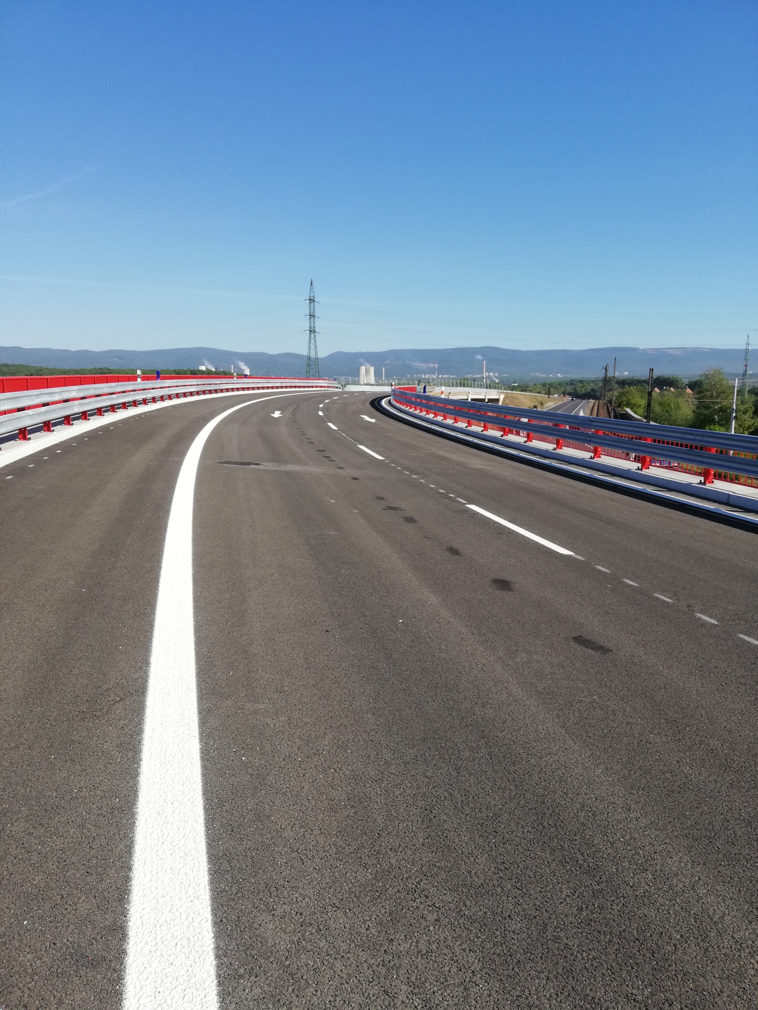 Nowa, asfaltowa droga z białymi pasami i czerwonymi barierkami, w tle słup wysokiego napięcia i zabudowania przemysłowe pod błękitnym niebem.