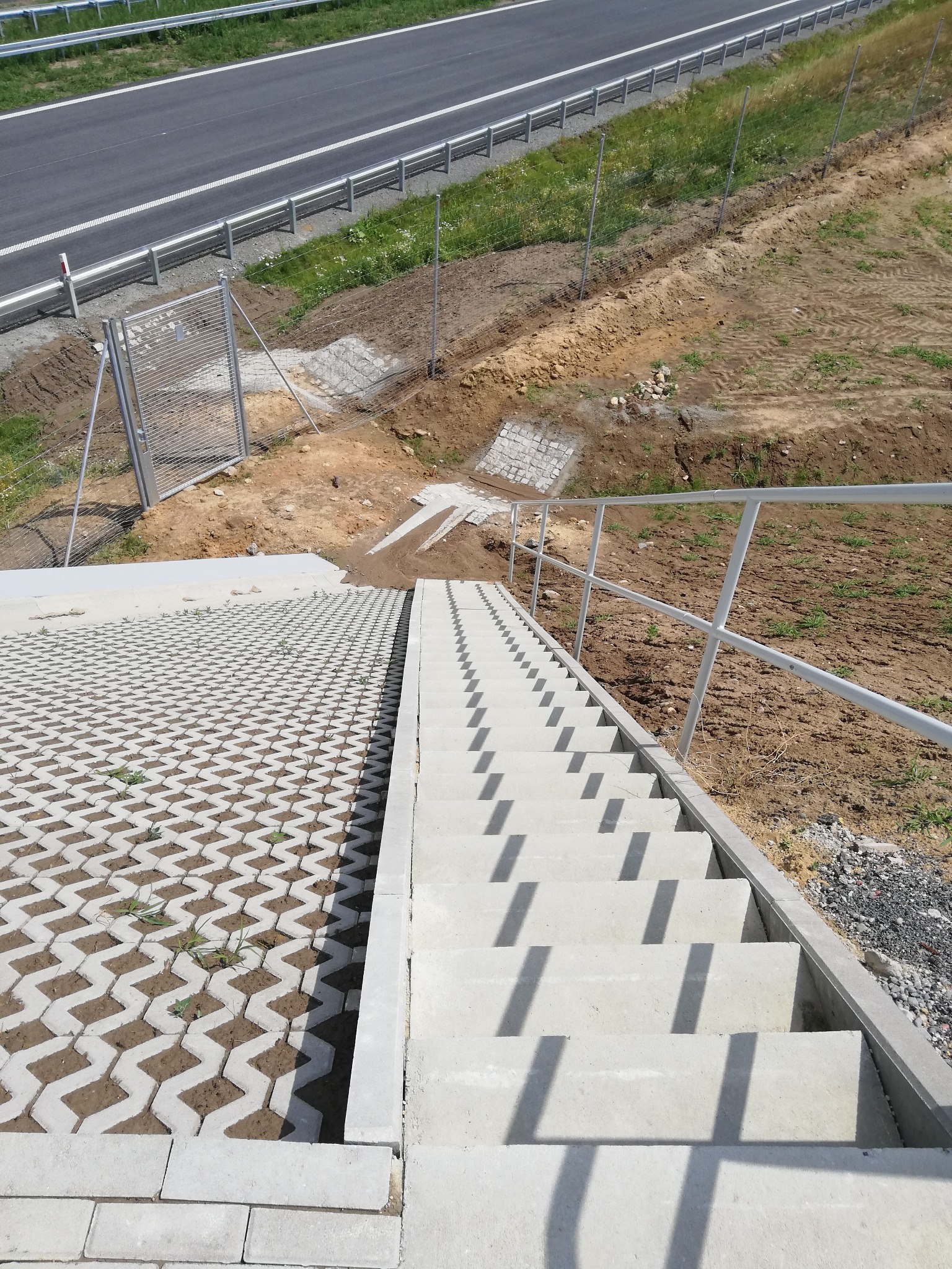 Schody betonowe prowadzące w dół, obok ażurowa nawierzchnia z kostki betonowej, widok z góry na nową drogę i ogrodzenie.