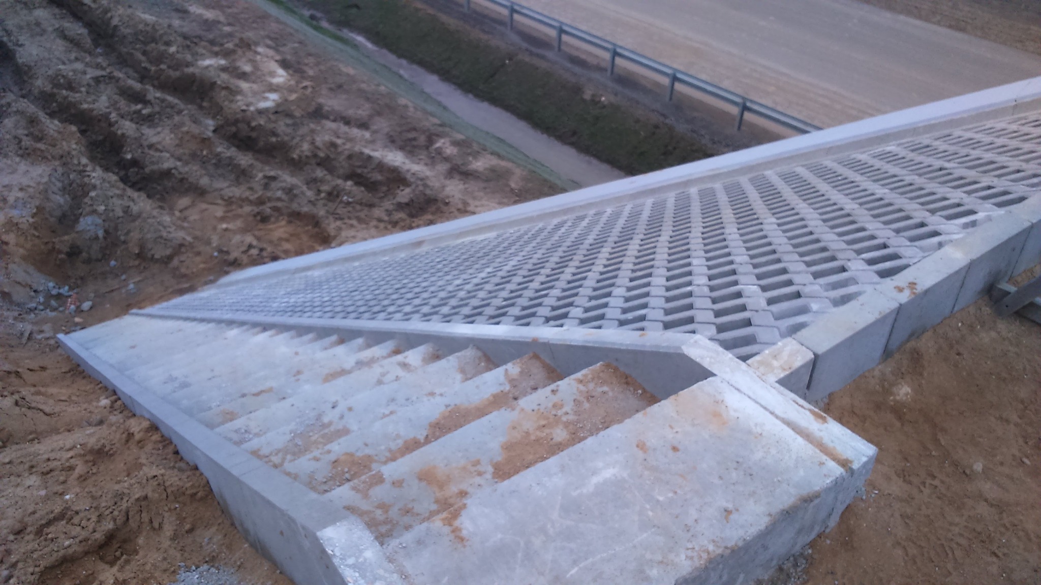 Betonowe schody z ażurową nawierzchnią na zboczu, widok z góry, z ziemią po bokach i drogą w tle.