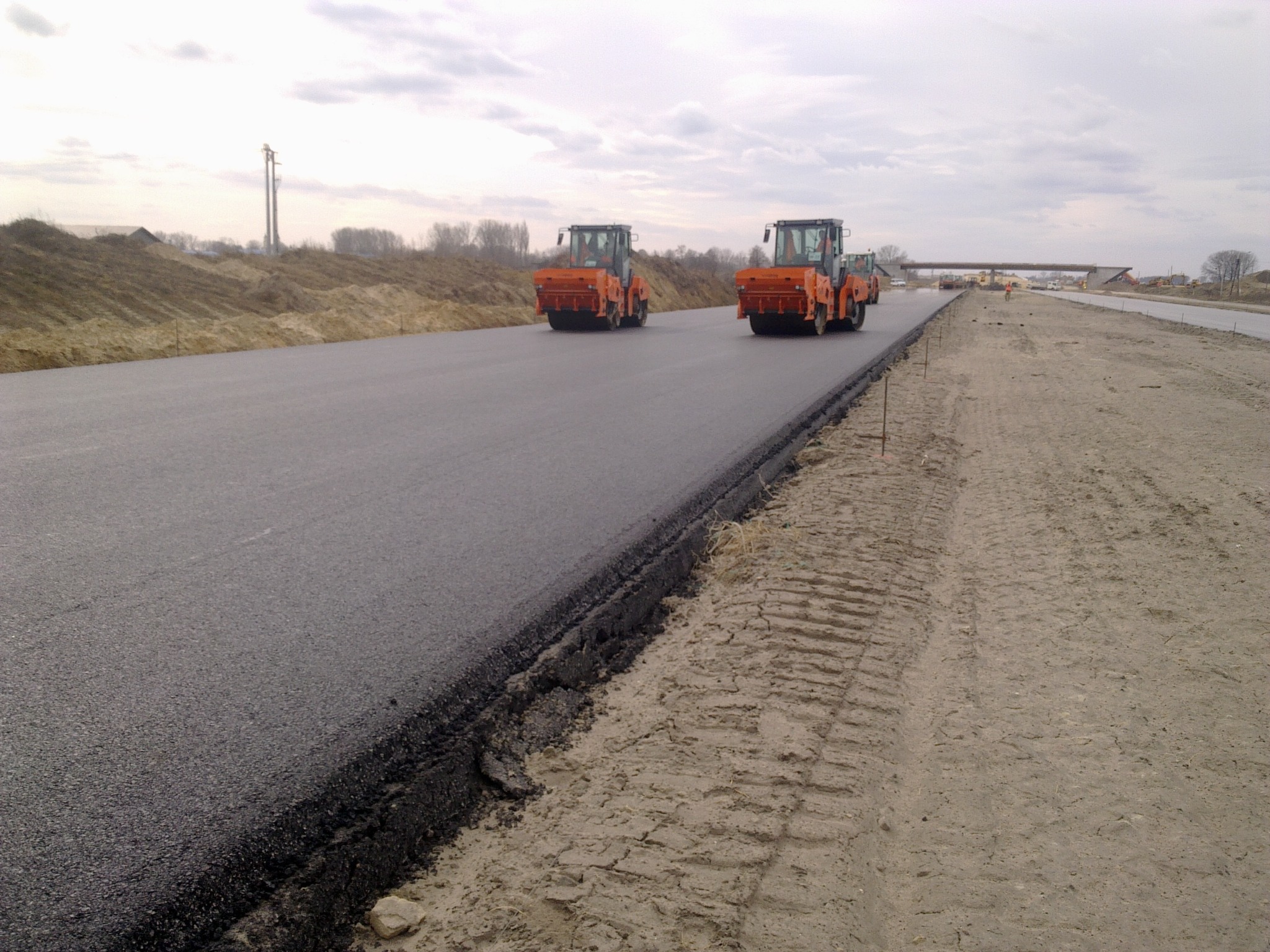 Dwa pomarańczowe walce drogowe jadą po świeżo wylanym asfalcie na budowie drogi, w tle widoczny wiadukt i piaszczyste pobocze.
