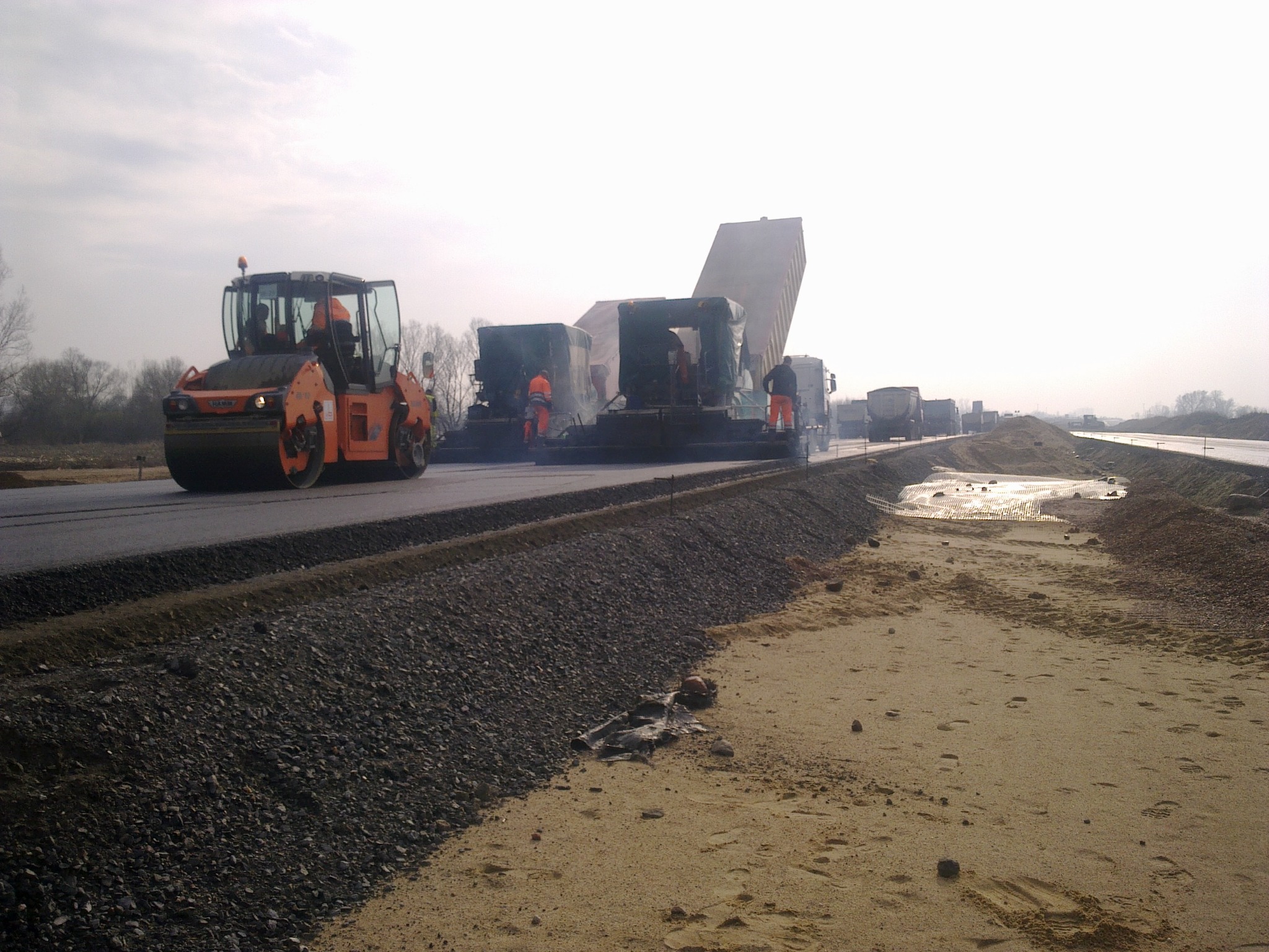 Walec drogowy ugniata świeżo położony asfalt na budowie drogi, w tle ekipa robotników obsługuje maszynę rozkładającą nawierzchnię, a za nią ustawia się kolejka ciężarówek z materiałem.