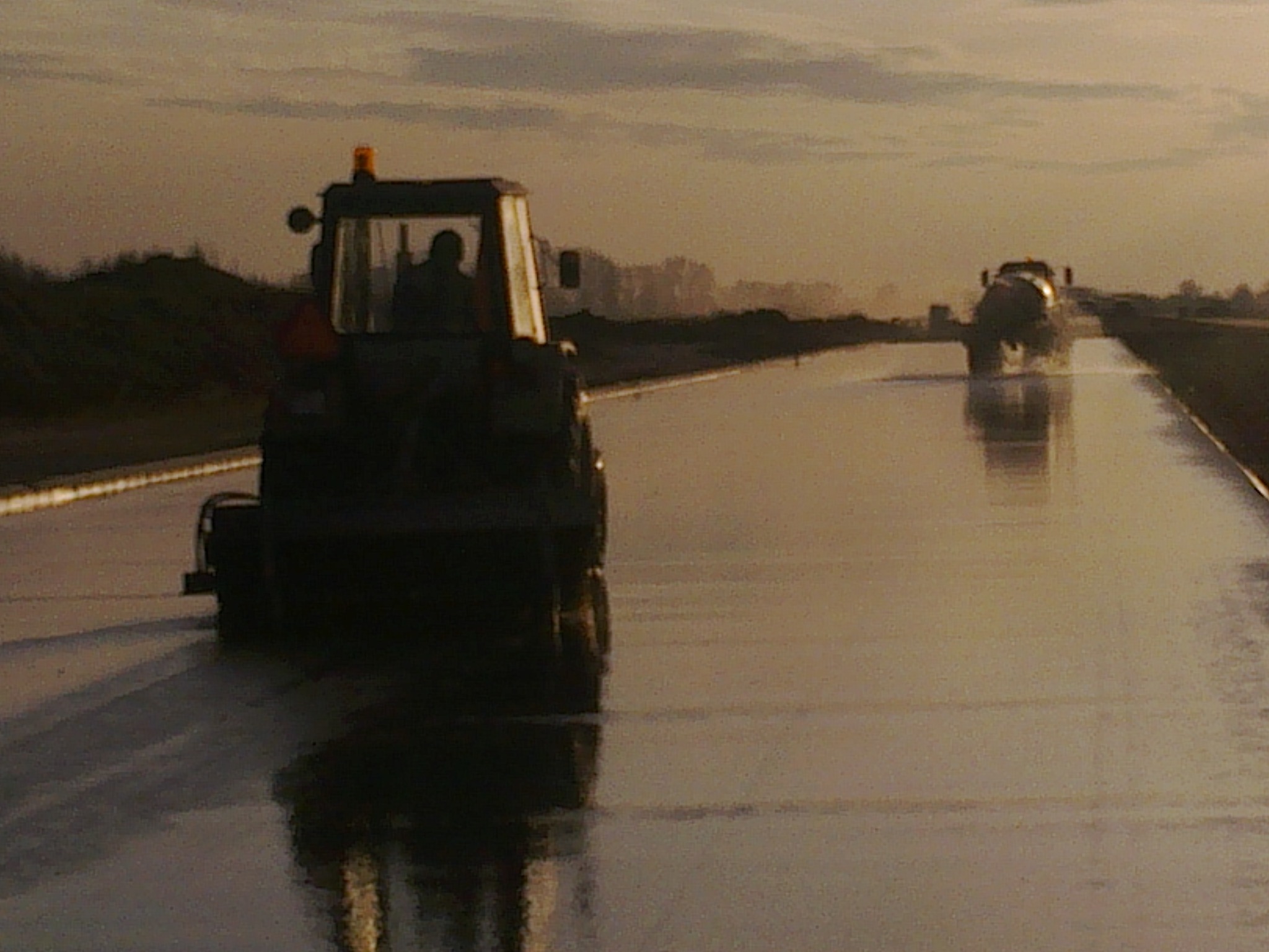 Dwa traktory z przyczepami pracują na mokrej, nowo wylanej drodze, odbijając zachodzące słońce.