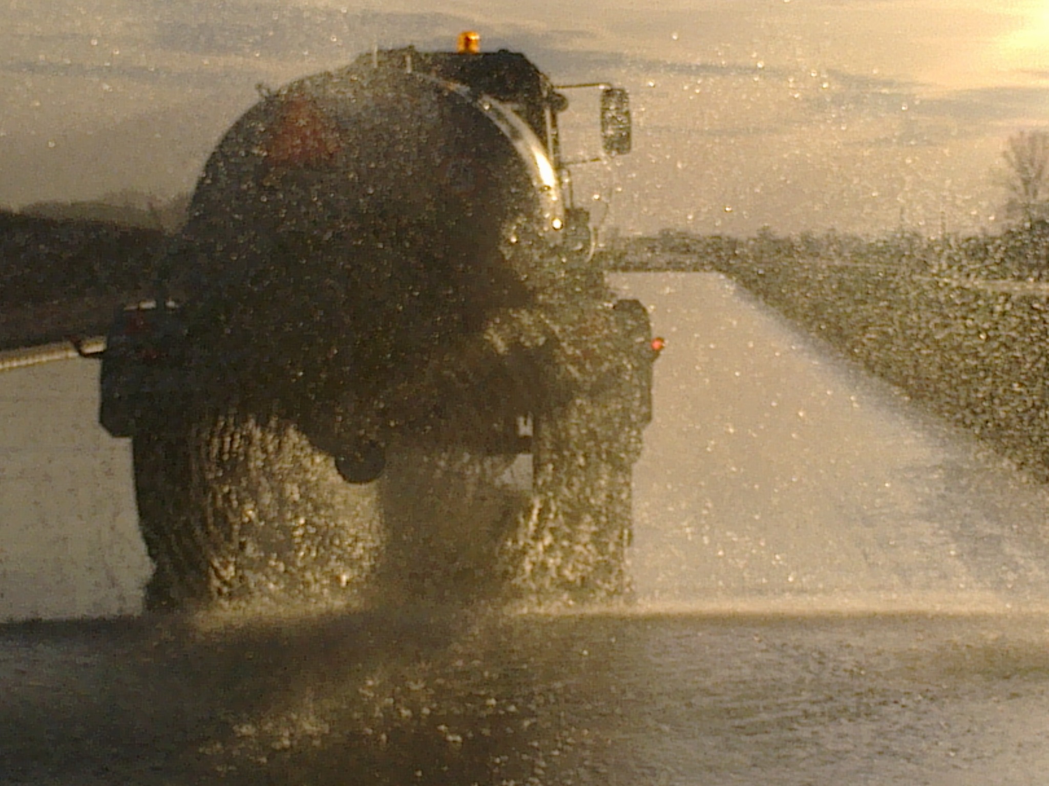 Ciężki pojazd z cysterną, rozpryskujący wodę na utwardzonej drodze, prawdopodobnie w celu jej utwardzenia lub zagęszczenia, podczas zachodu słońca.