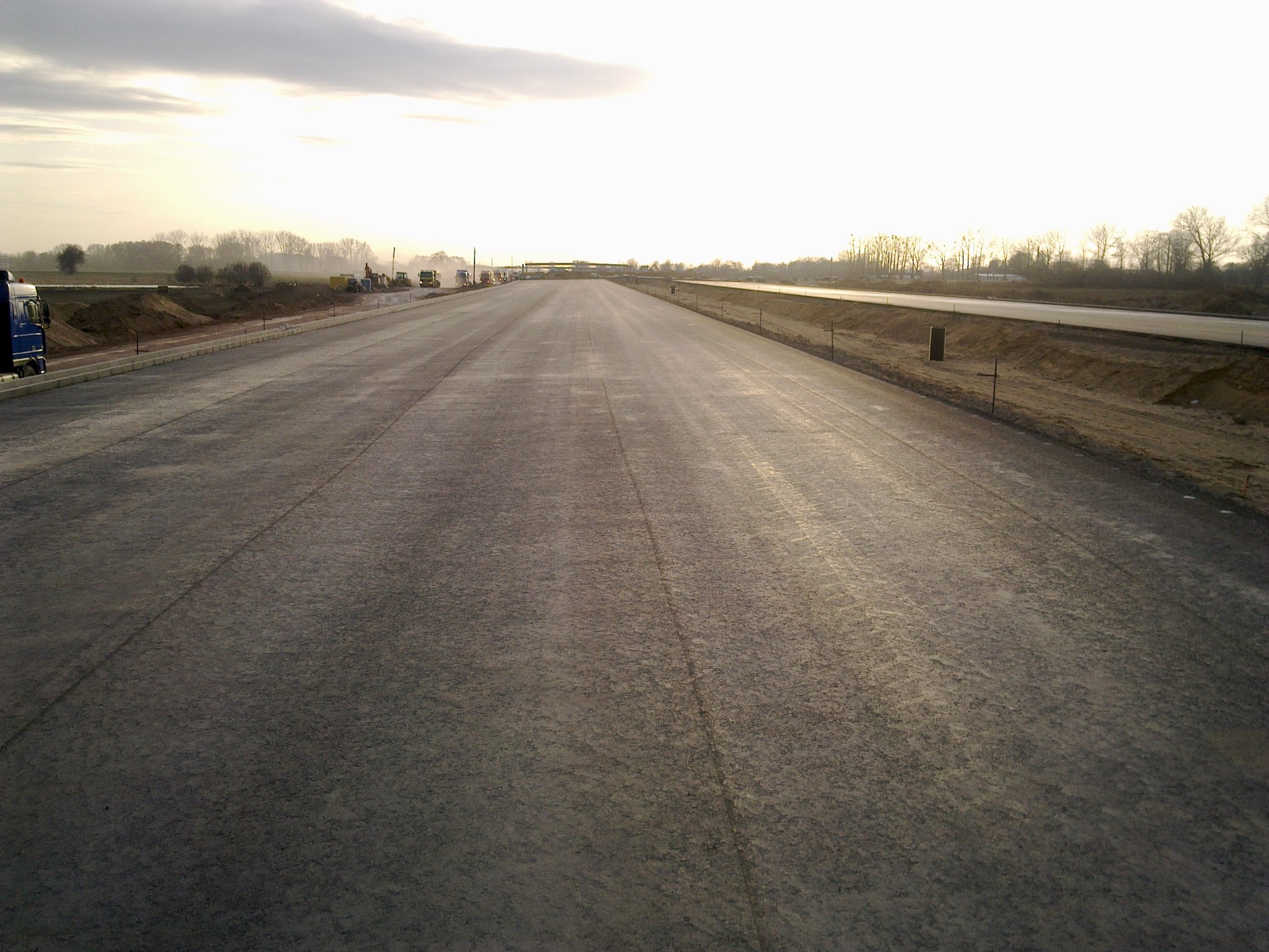 Szeroka droga w trakcie budowy, z widocznymi maszynami budowlanymi w oddali, świeżo wylany asfalt, przygotowany do dalszych prac wykończeniowych, w tle drzewa i pochmurne niebo.