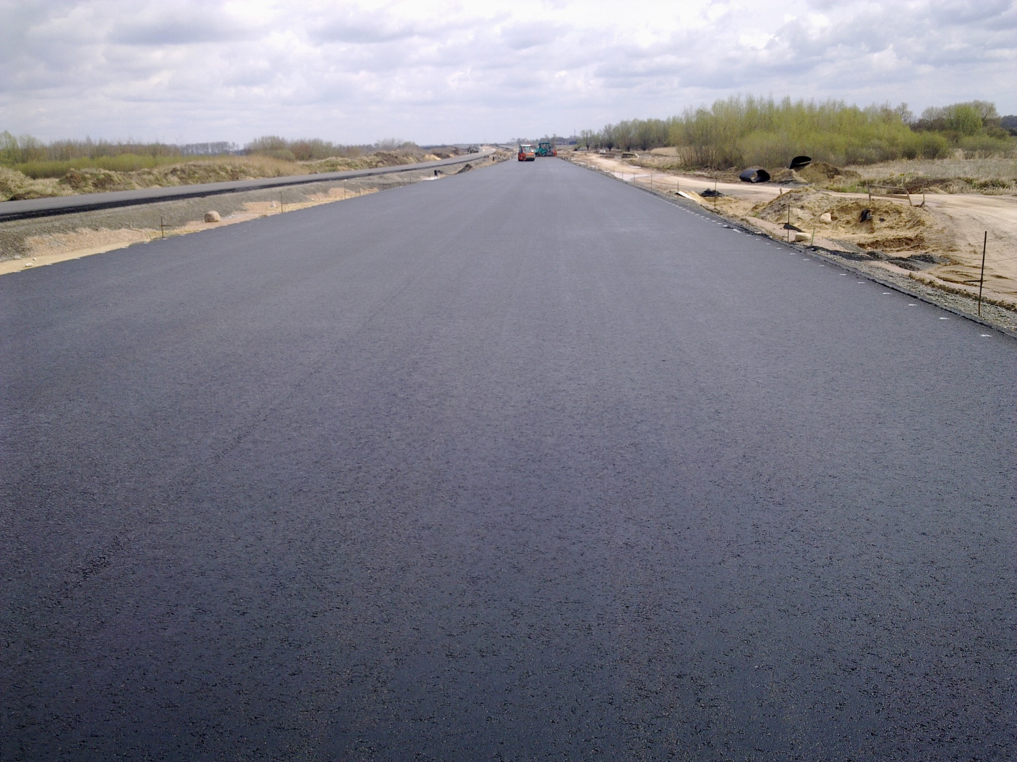 Nowo położona, gładka nawierzchnia asfaltowa drogi w trakcie budowy, z widocznymi maszynami budowlanymi w oddali i elementami przygotowania terenu po bokach.
