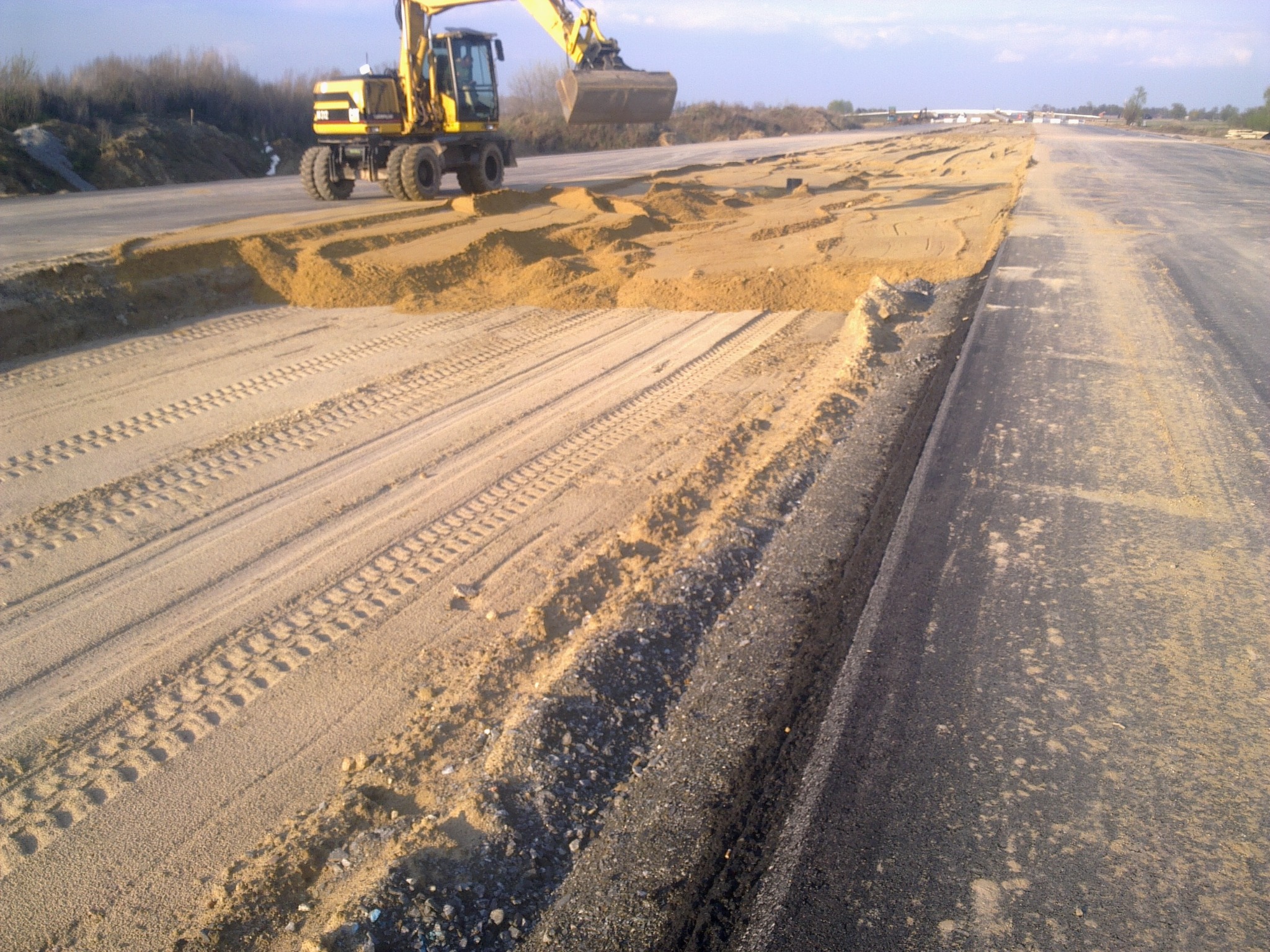 Żółta koparka kołowa rozprowadza piasek na przygotowywanym podłożu drogi, widoczne ślady opon na piasku, asfaltowa nawierzchnia z prawej strony.