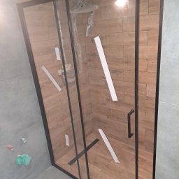Igor tam - Kabina prysznicowa z czarnymi profilami, zamontowana w łazience z szarymi i drewnopodobnymi płytkami, zabezpieczona taśmą malarską w trakcie prac wykończeniowych.