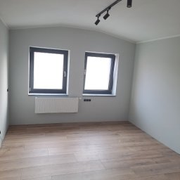 Igor tam - Pusty pokój po remoncie z panelami podłogowymi w odcieniach brązu, szare ściany, dwa okna z czarnymi ramami, biały grzejnik pod oknem i czarna listwa z czterema reflektorami na suficie.